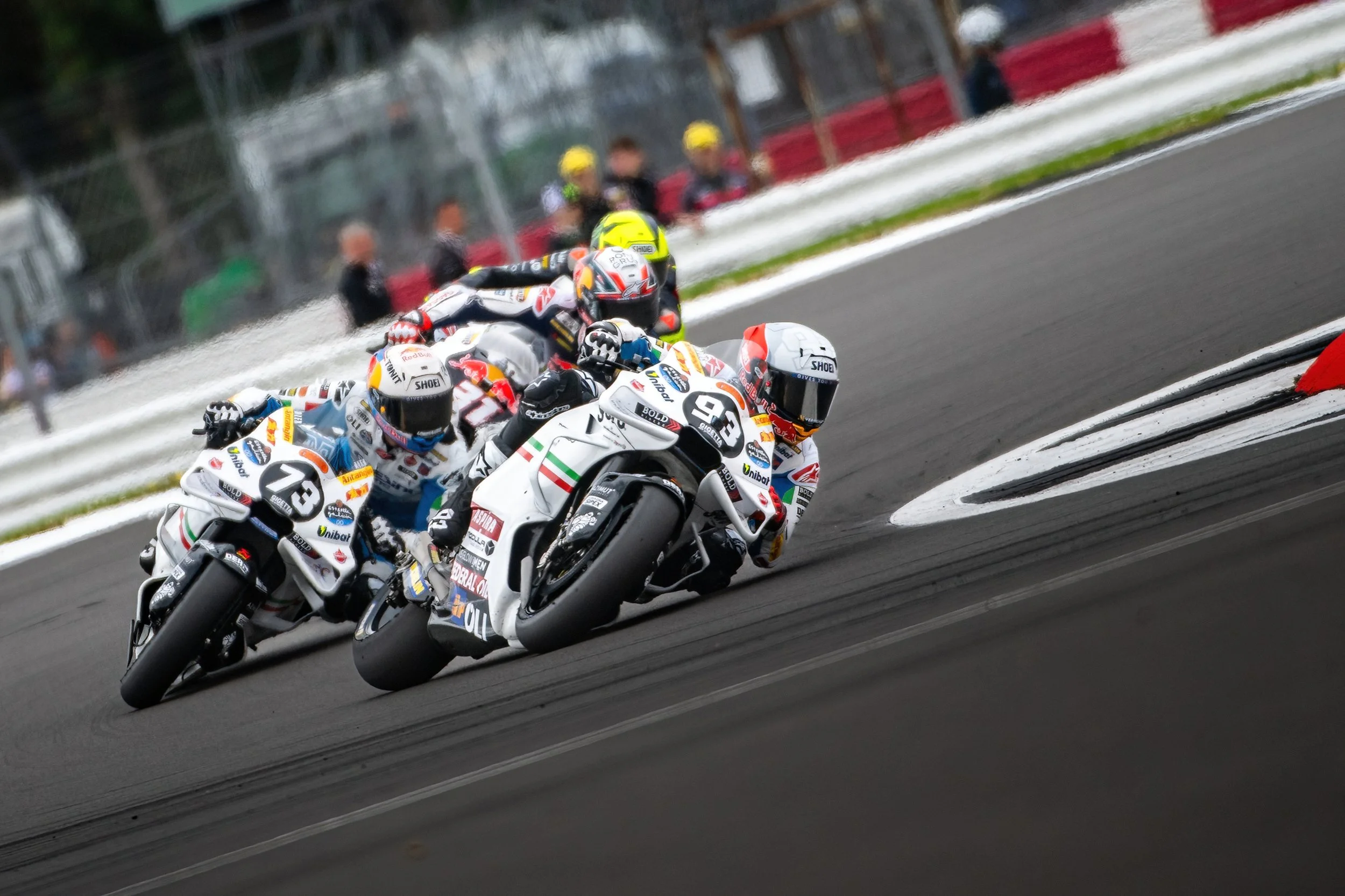 Motorcycle racers leaning into a turn on a race track, with the leading rider in a white suit and helmet, followed by three racers behind him. Spectators and racers are visible in the background.