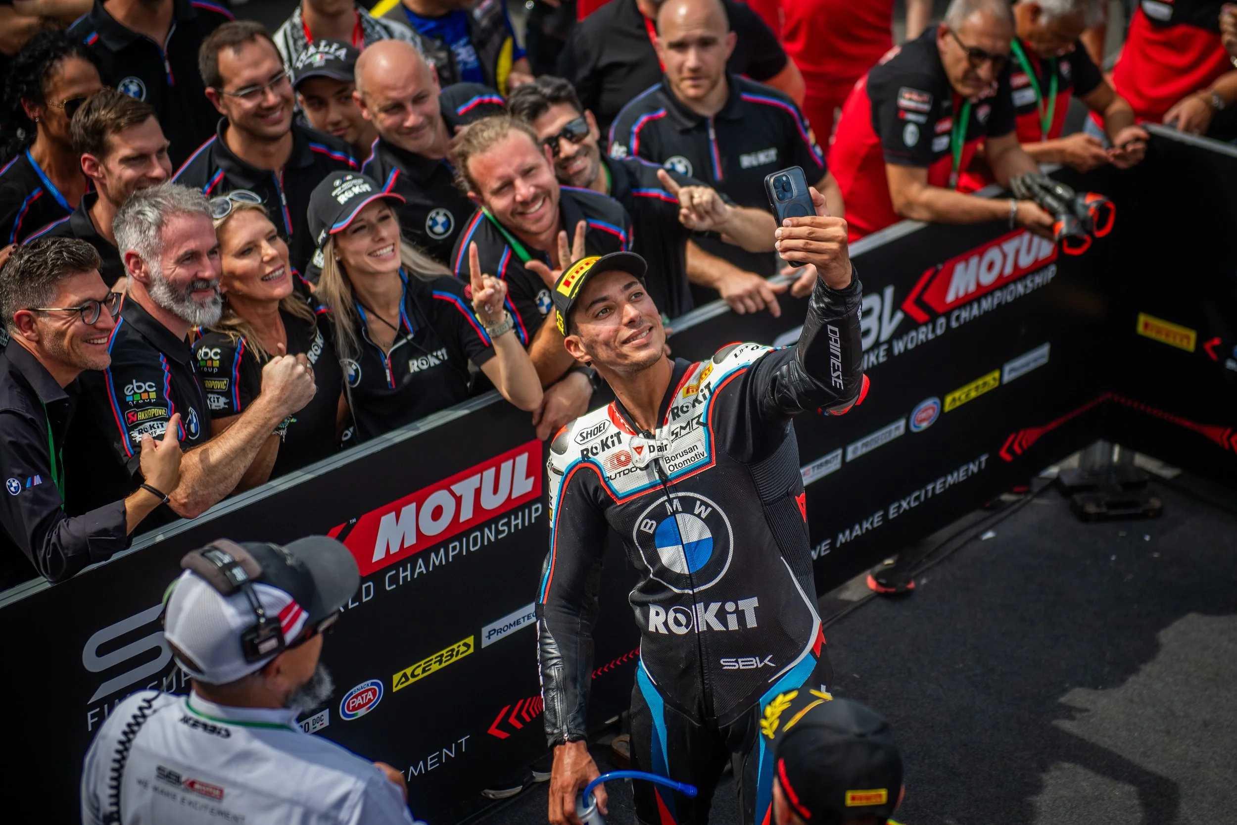Race car driver taking a selfie with cheering fans and team members at a motorsport event.