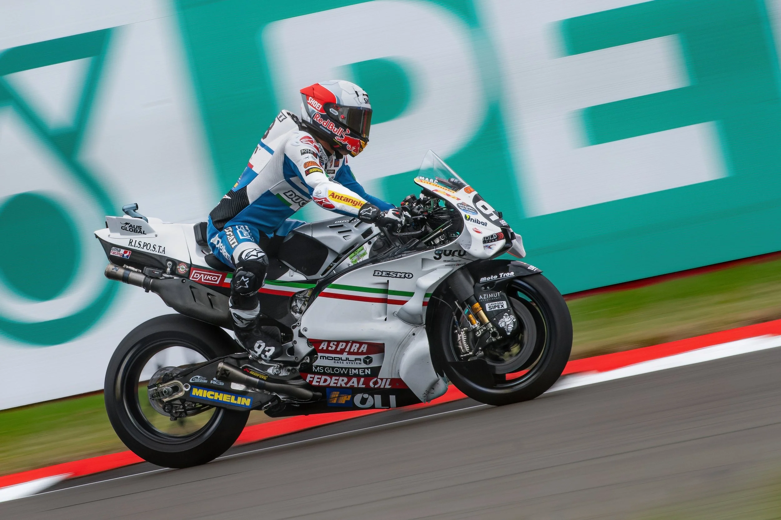 A professional motorcycle racer in a blue, white, and black suit riding a white racing motorcycle on a track with a red and white curb, against a blurred green background.