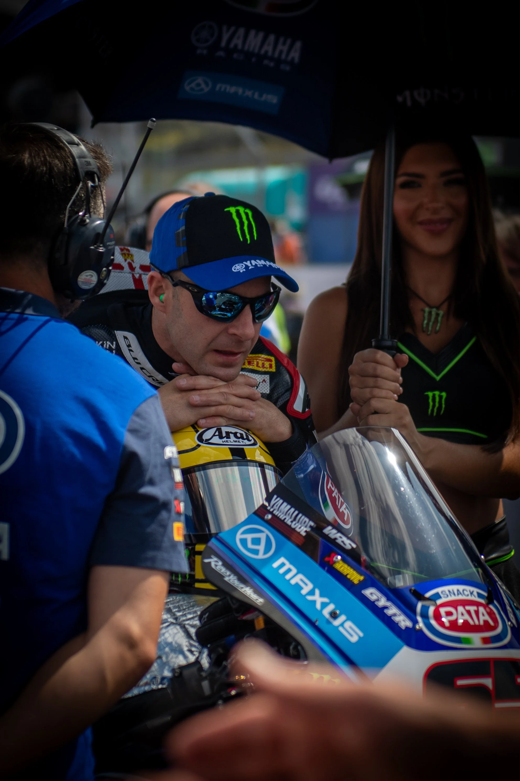 A professional motorcycle racer with a yellow helmet, wearing sunglasses and a racing suit, is in a paddock area surrounded by team members and a woman holding an umbrella with the Monster Energy logo. The motorcycle has sponsor logos including Yamah