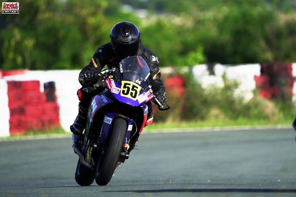 A motorcycle racer in black gear and helmet leaning into a turn on a racetrack, with the number 55 on the motorcycle and blurred green foliage in the background. rider name Lawrence Valencia