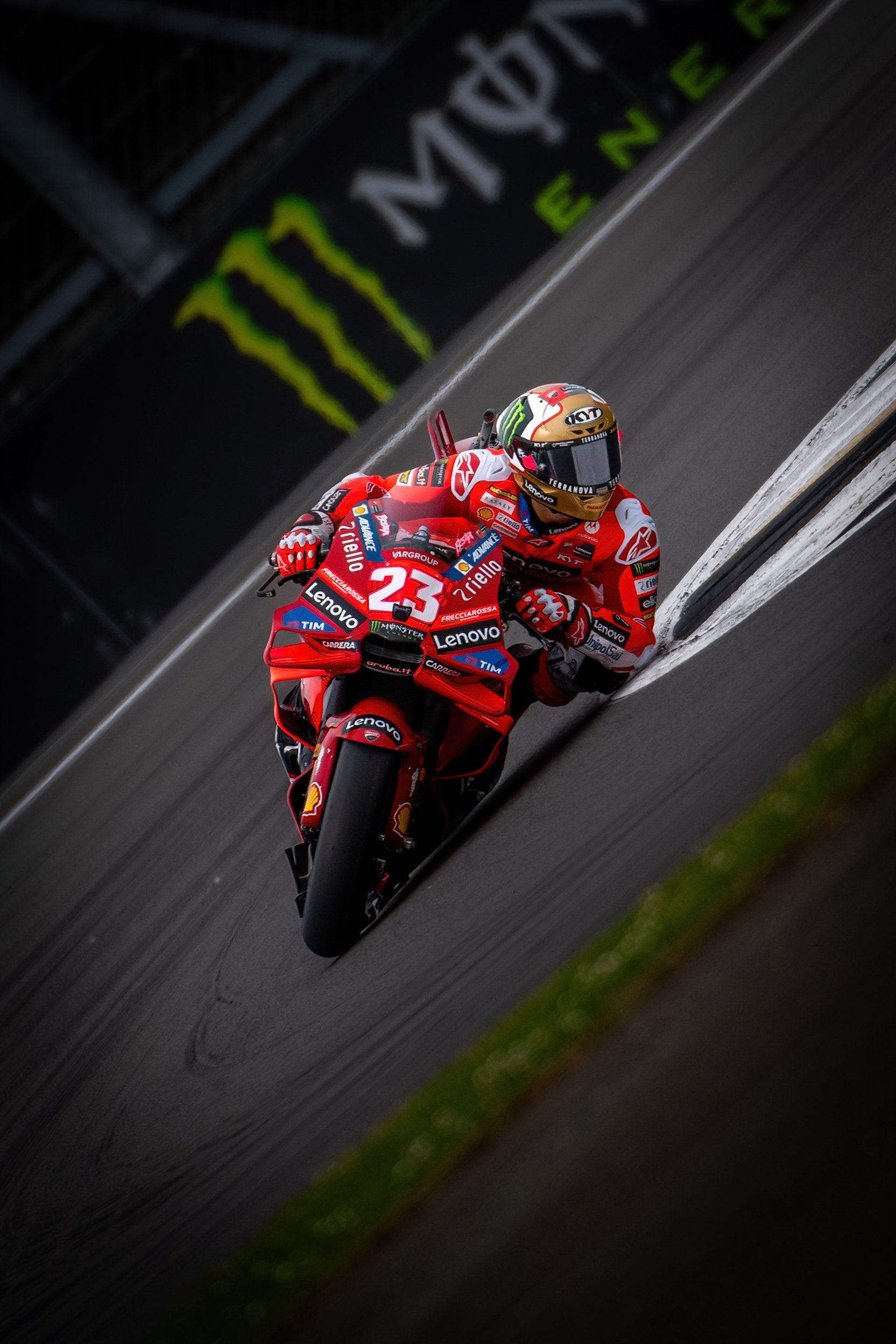 Motorcycle racer in red riding gear and helmet on a red Ducati racing bike, leaning into a turn on a racetrack with Monster Energy signs in the background.