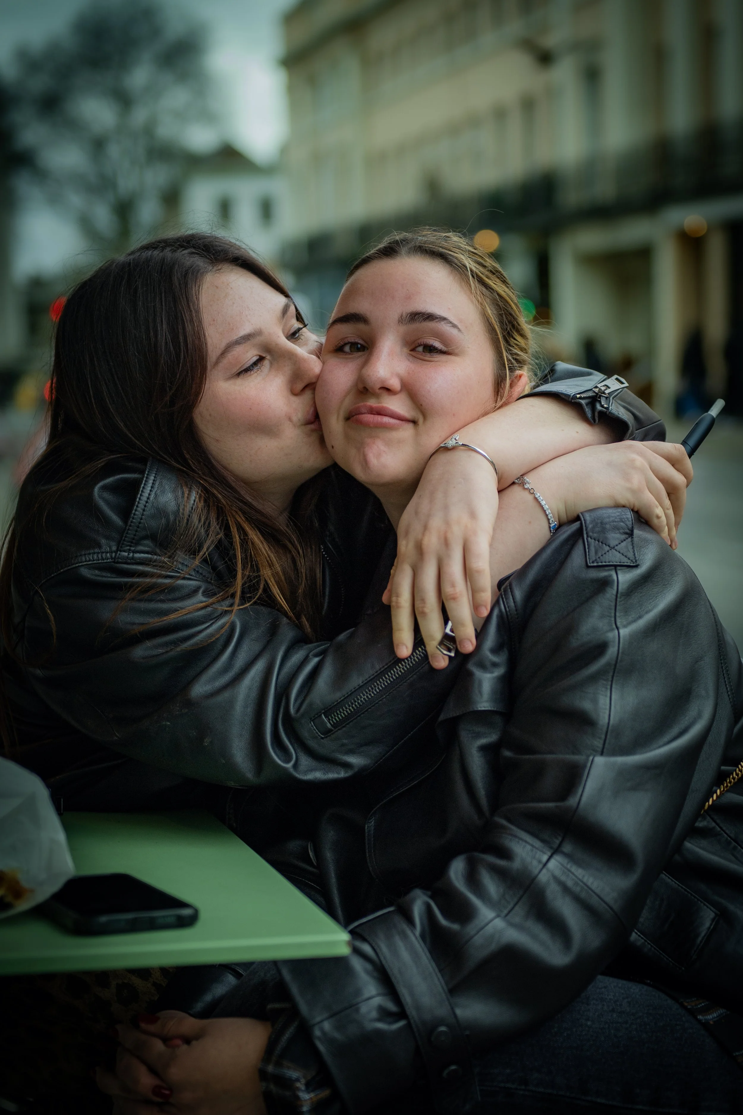 Two women, one with dark hair giving a kiss on the cheek to the other with lighter hair, in an outdoor urban setting.