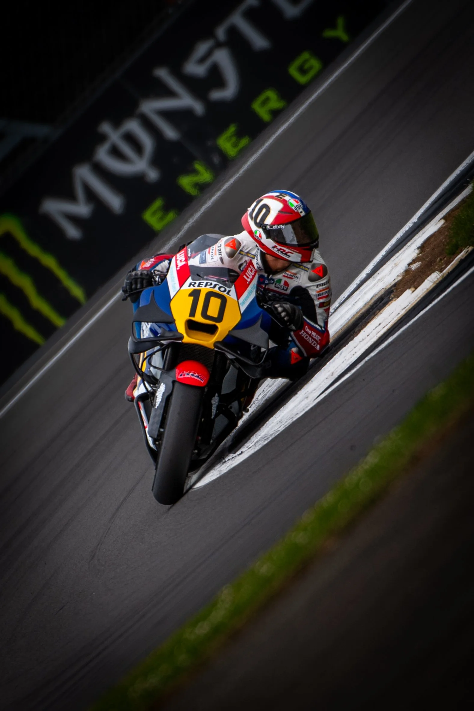 Motorcycle racer leaning into a turn on a race track, wearing a helmet and racing suit, with a yellow number 10 on the front of the bike and a background featuring a billboard with the Monster Energy logo.