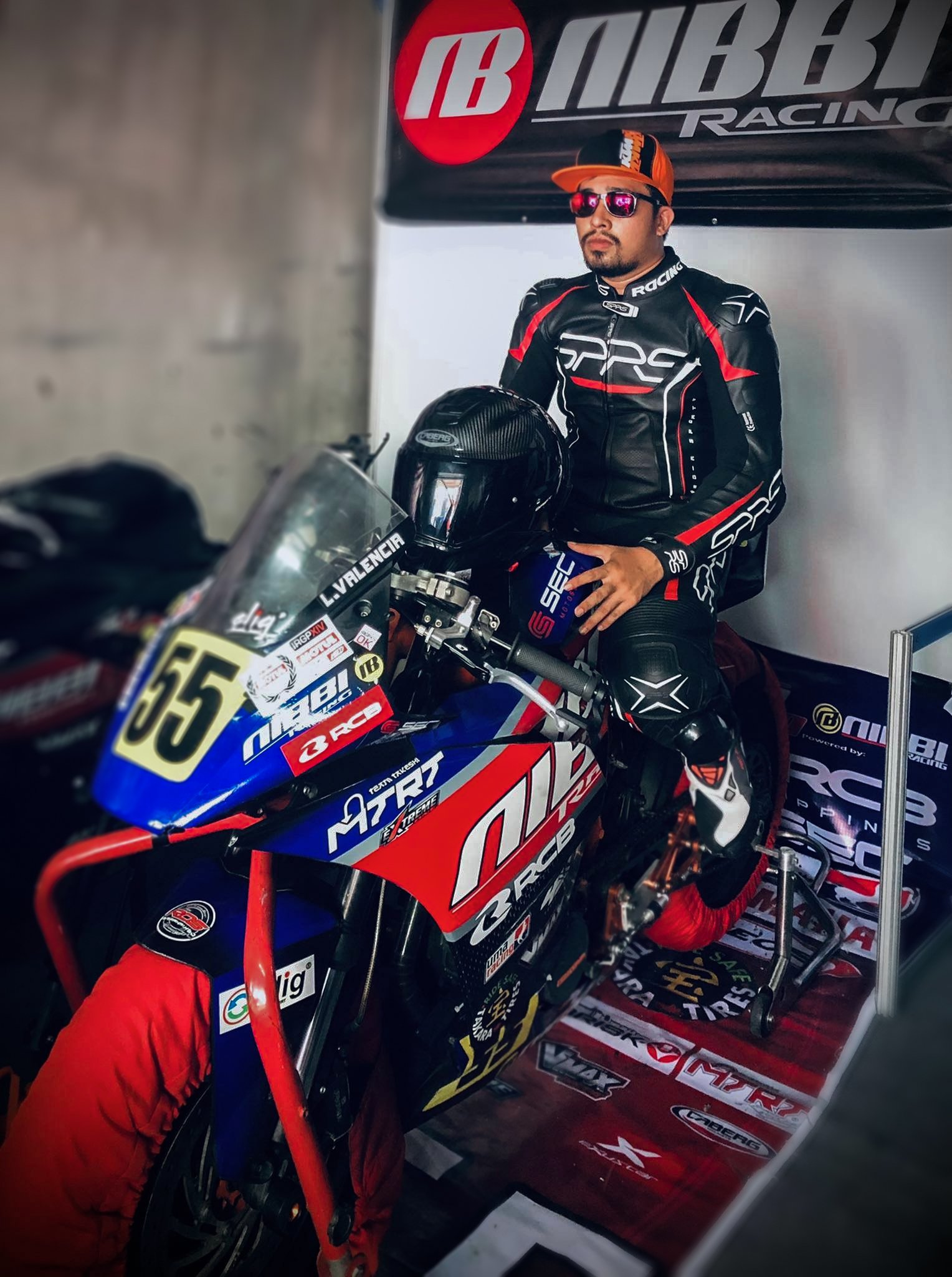 Lawrence Valencia, wearing a black racing suit with red and white accents, sunglasses, and a cap, with a ride number 55 on the motorcycle.