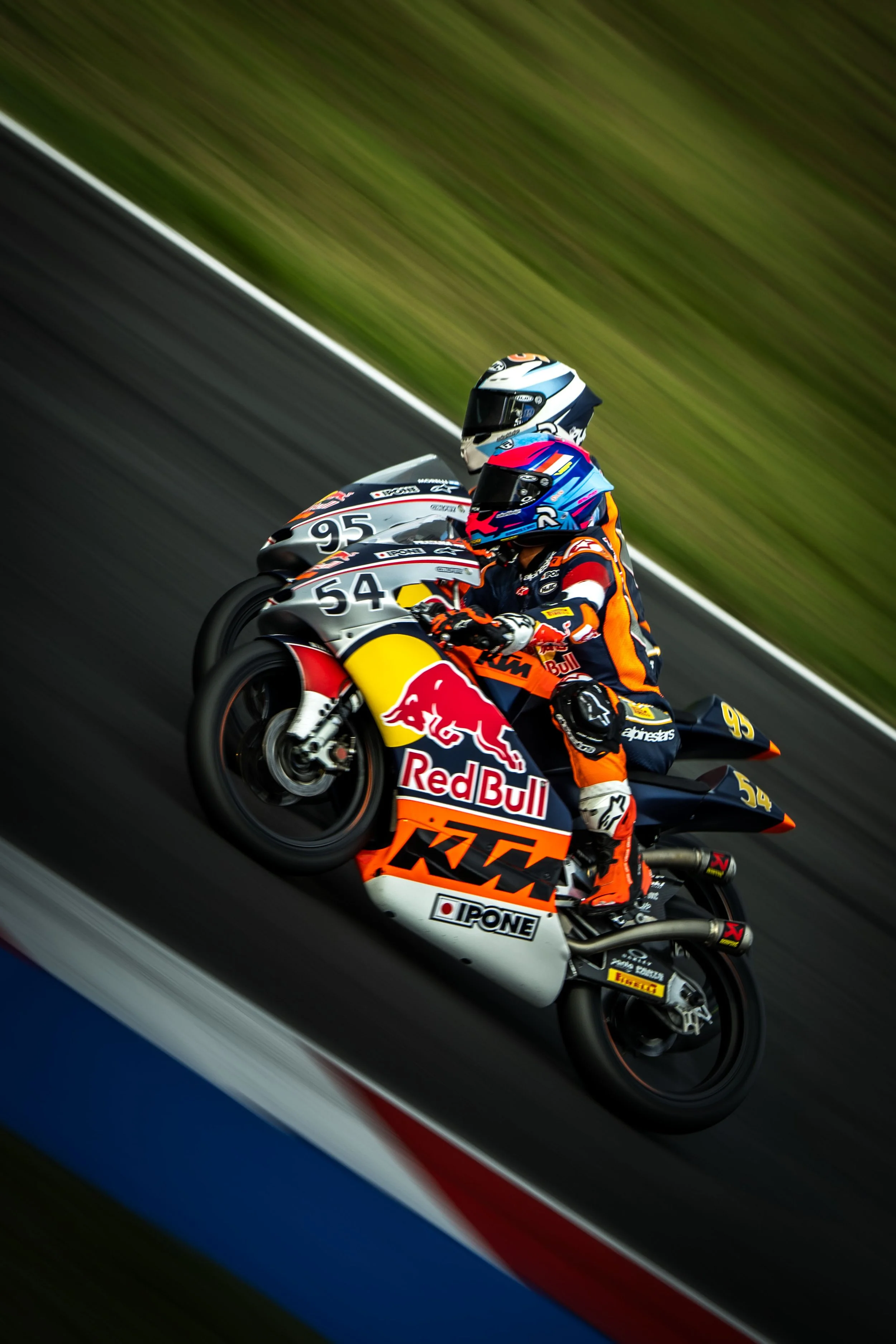 Two motorcycle racers in full gear leaning into a turn on a racetrack, sponsored by Red Bull and KTM.