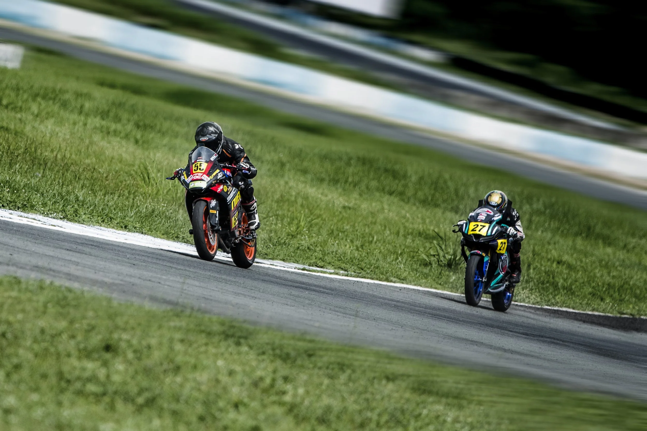 Lawrence Valencia and JD Purino racing on a track, leaning into a curve with grass on the side and blurred background.