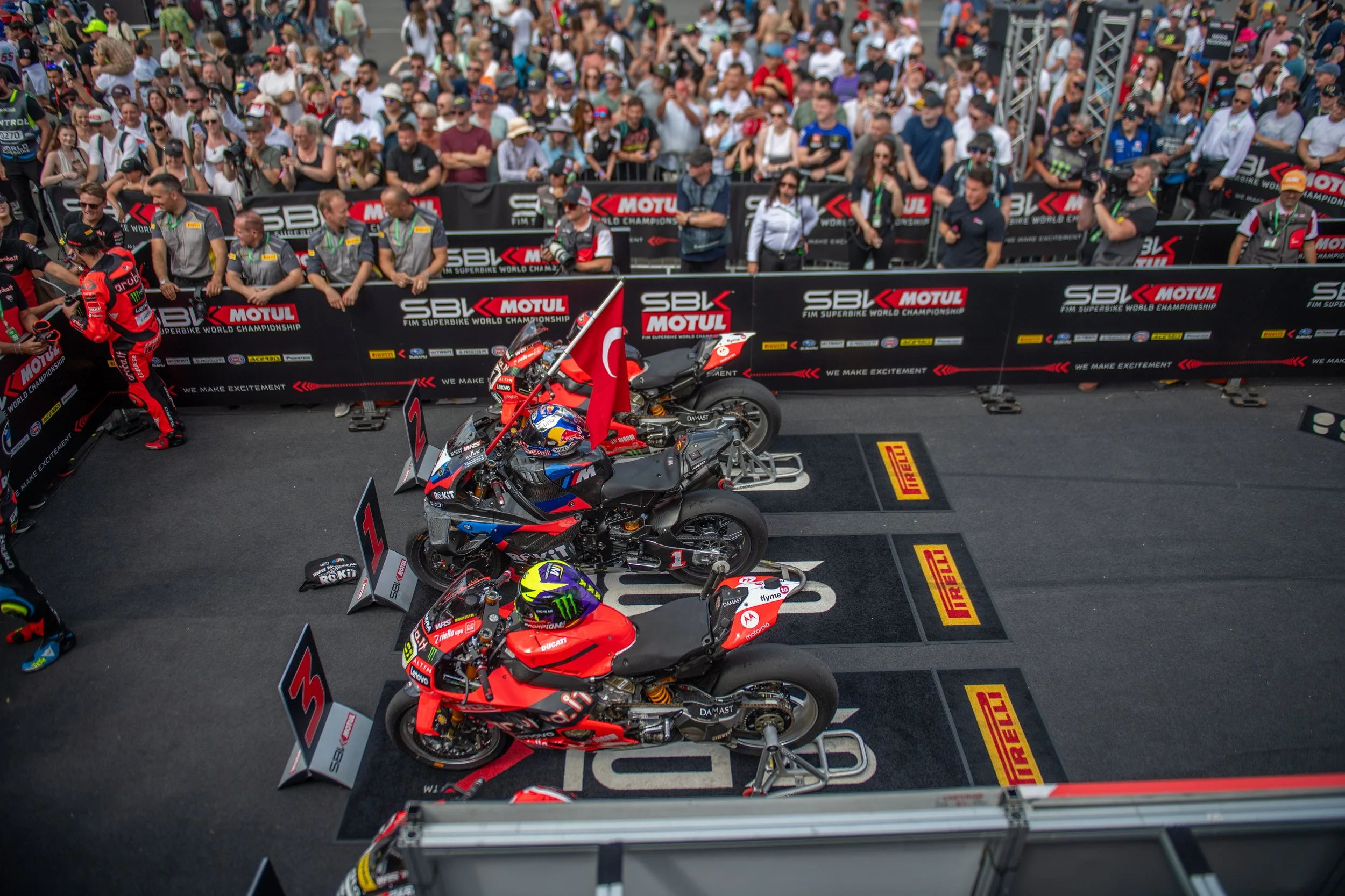 Motorcycle racing event with three racing bikes on a podium, crowds of spectators, and team members behind barriers at a Superbike World Championship race.