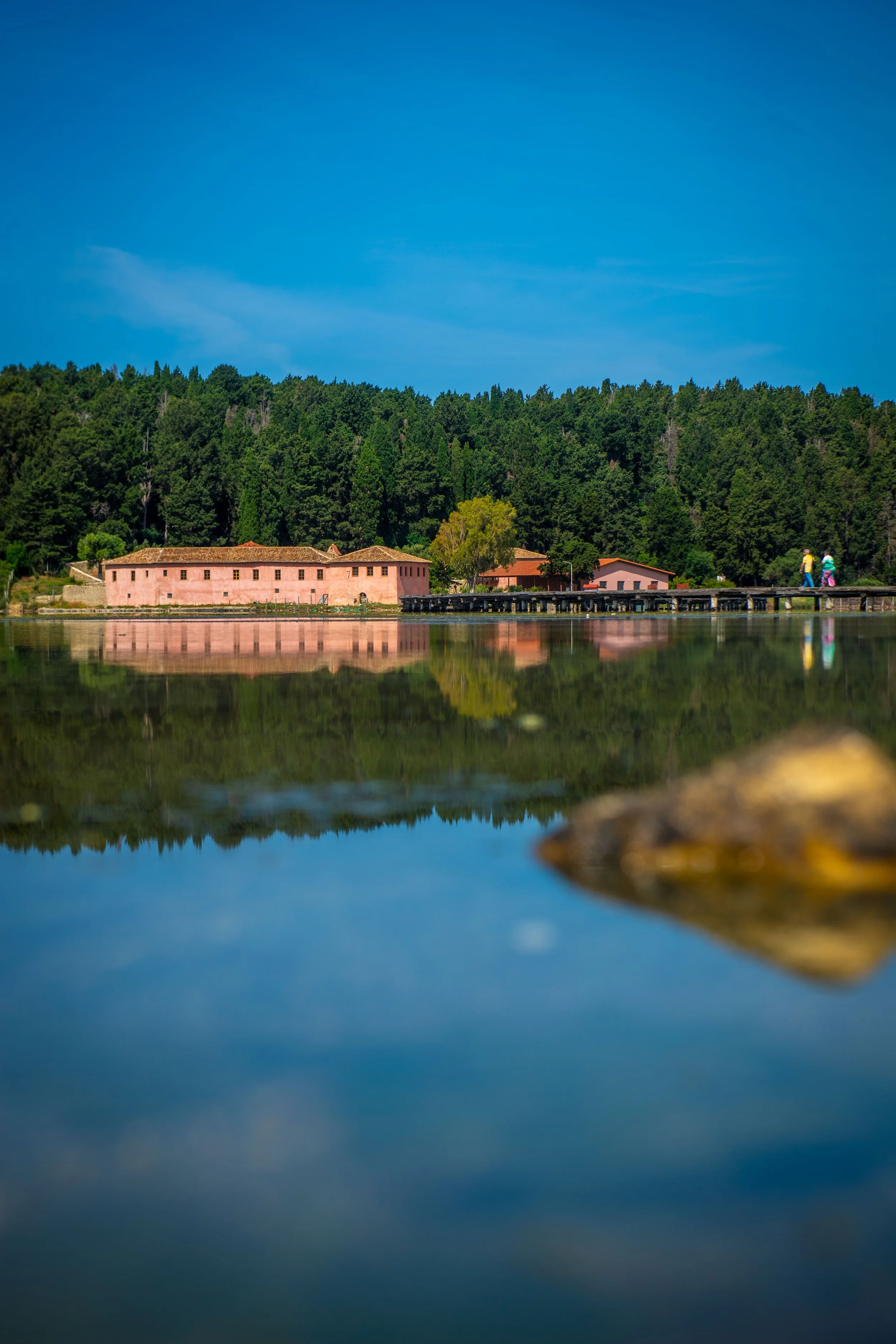 A lakeside scene with a pink building and smaller structures, surrounded by dense green trees under a blue sky, with a dock and three people walking on it.