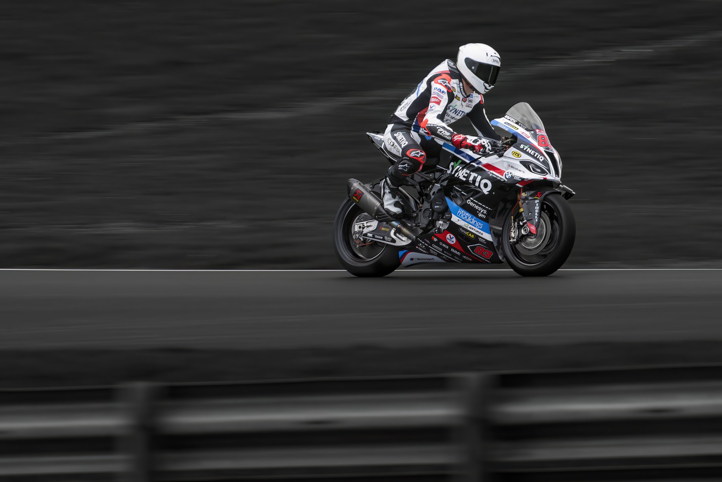 A motorcyclist in racing gear riding a sports bike on a race track, with dark blurred background and safety barriers in the foreground.