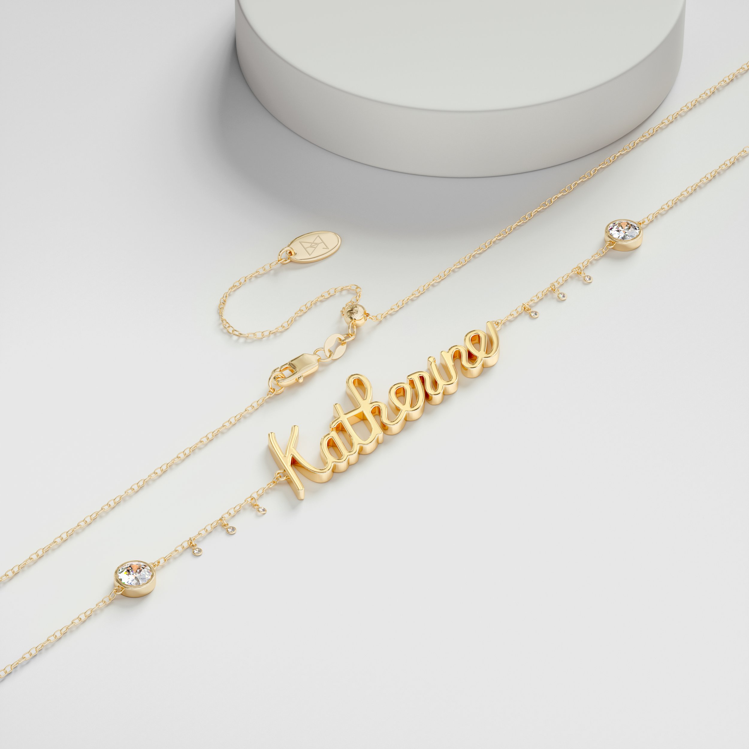 Gold jewelry necklace with the name 'katherine' in cursive and small dangling charms, alongside a delicate chain with small stones, all on a white surface.