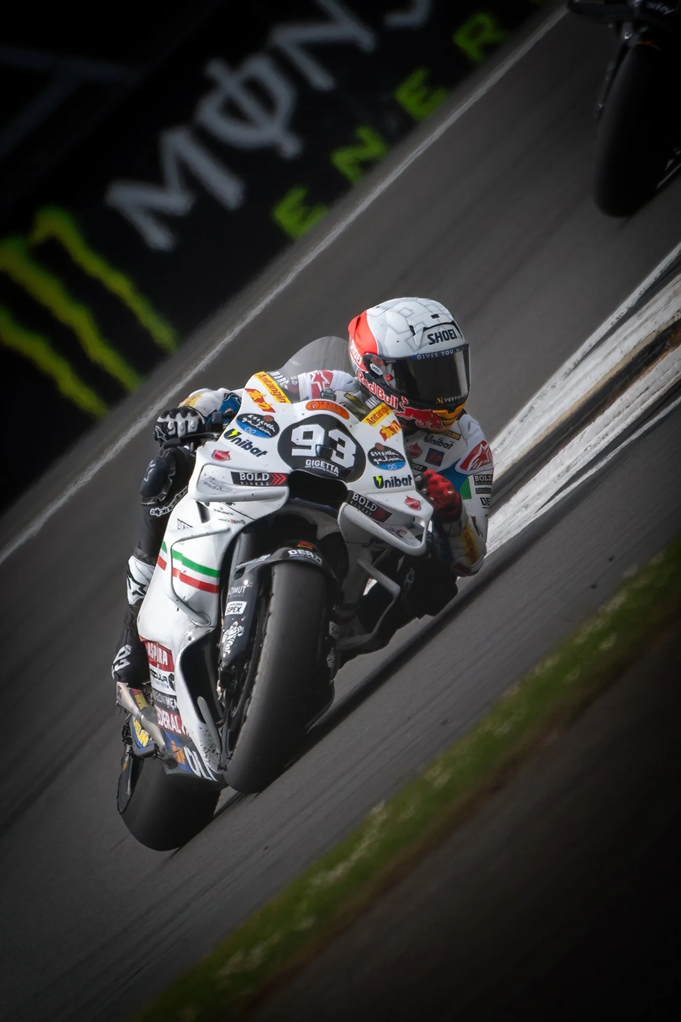 Motorcycle racer in full gear riding on a race track, leaning into a turn. The bike is white with various sponsor stickers, and the rider's helmet is white with a red and black design.