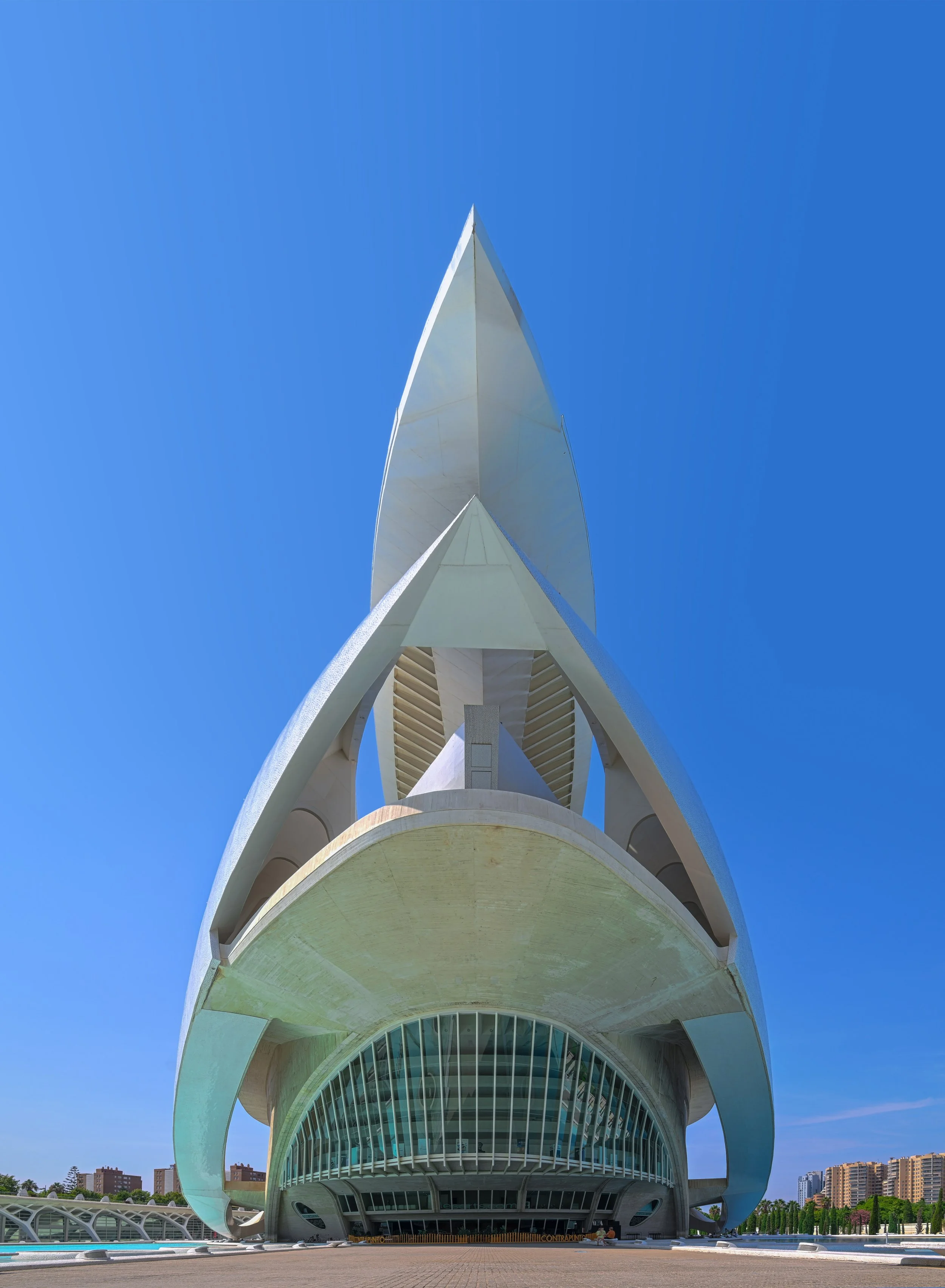A modern, white architectural building with a pointed, sail-like structure against a clear blue sky. The building features curved lines, large glass windows at the base, and an intricate design.