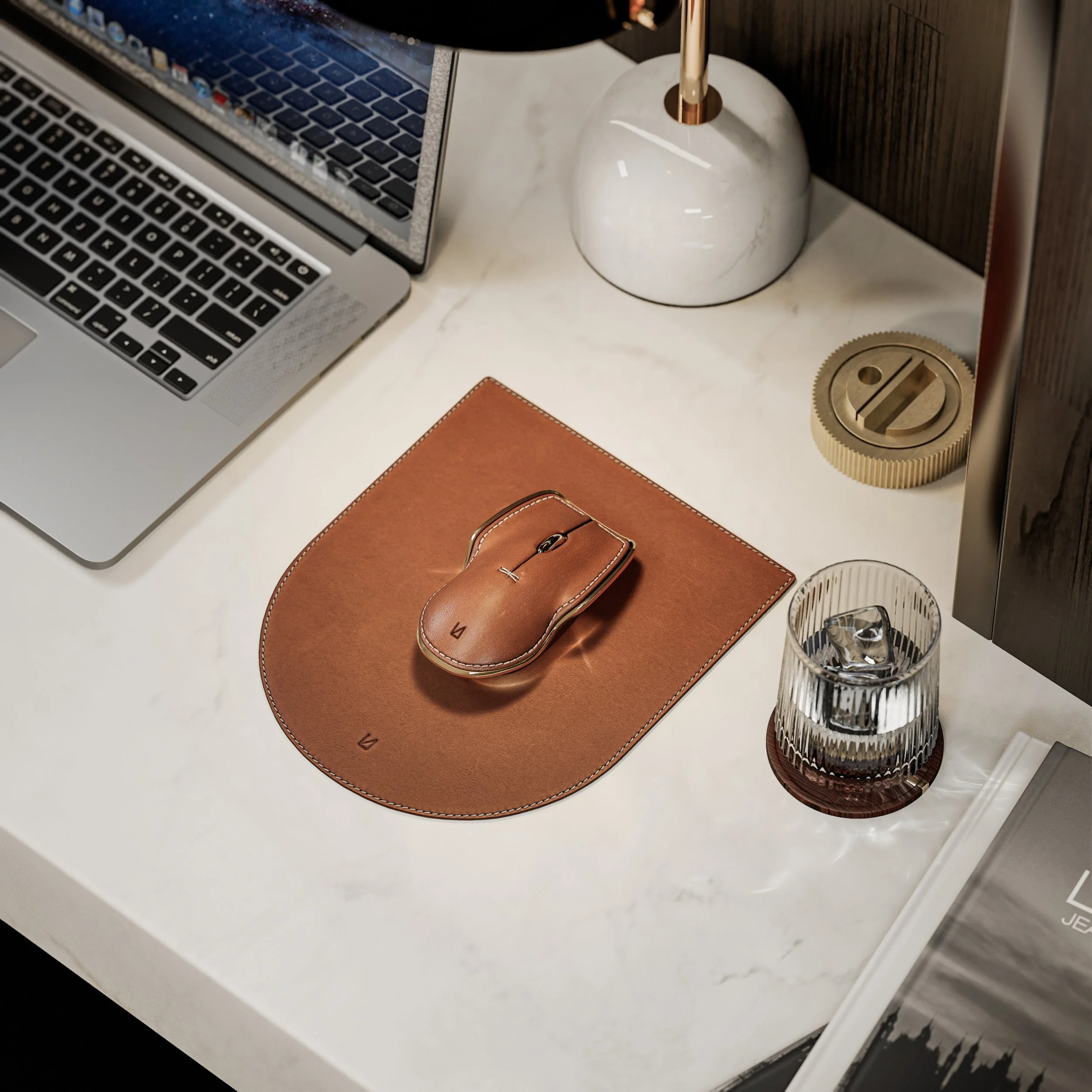 A workspace with a silver MacBook laptop, a white marble vase with a gold-colored rod, a round gold-colored object, a glass of water with ice on a leather coaster, and a leather mouse pad with a matching leather Lunar Artefacts mouse placed on it.