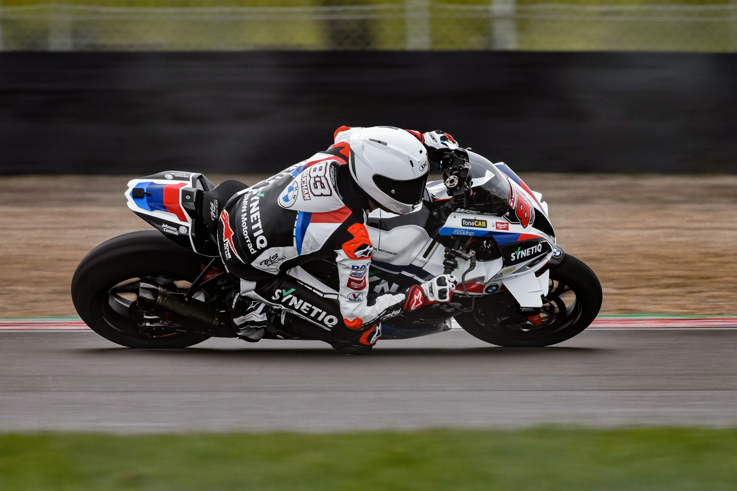 Motorcycle racer leaning into a turn on a racetrack, wearing full gear and a helmet, with the motorcycle featuring a BMW and Synetio sponsorship.