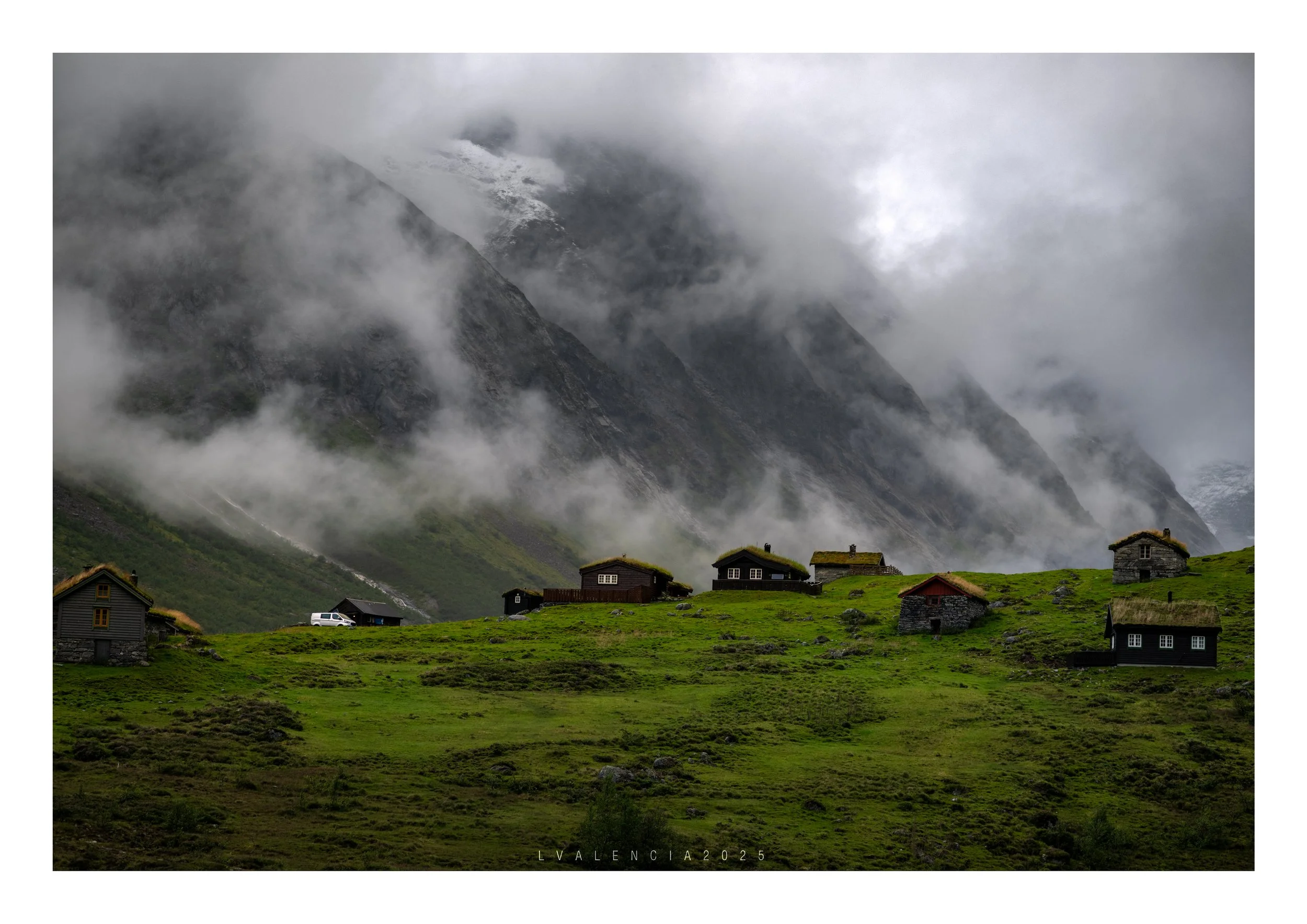 A green hillside with multiple small, dark-colored houses and a white vehicle, set against a backdrop of tall, misty mountains with clouds partially covering them.