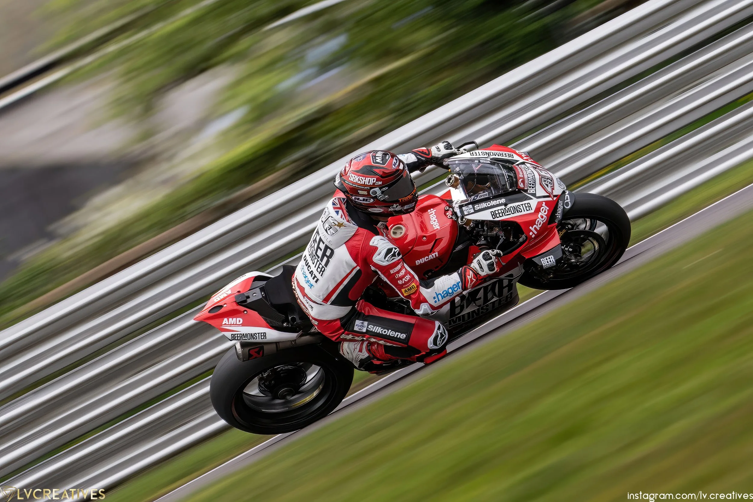 A motorcycle racer in red and white riding gear on a red Ducati motorcycle on a race track with blurred green grass and barriers in the background.
