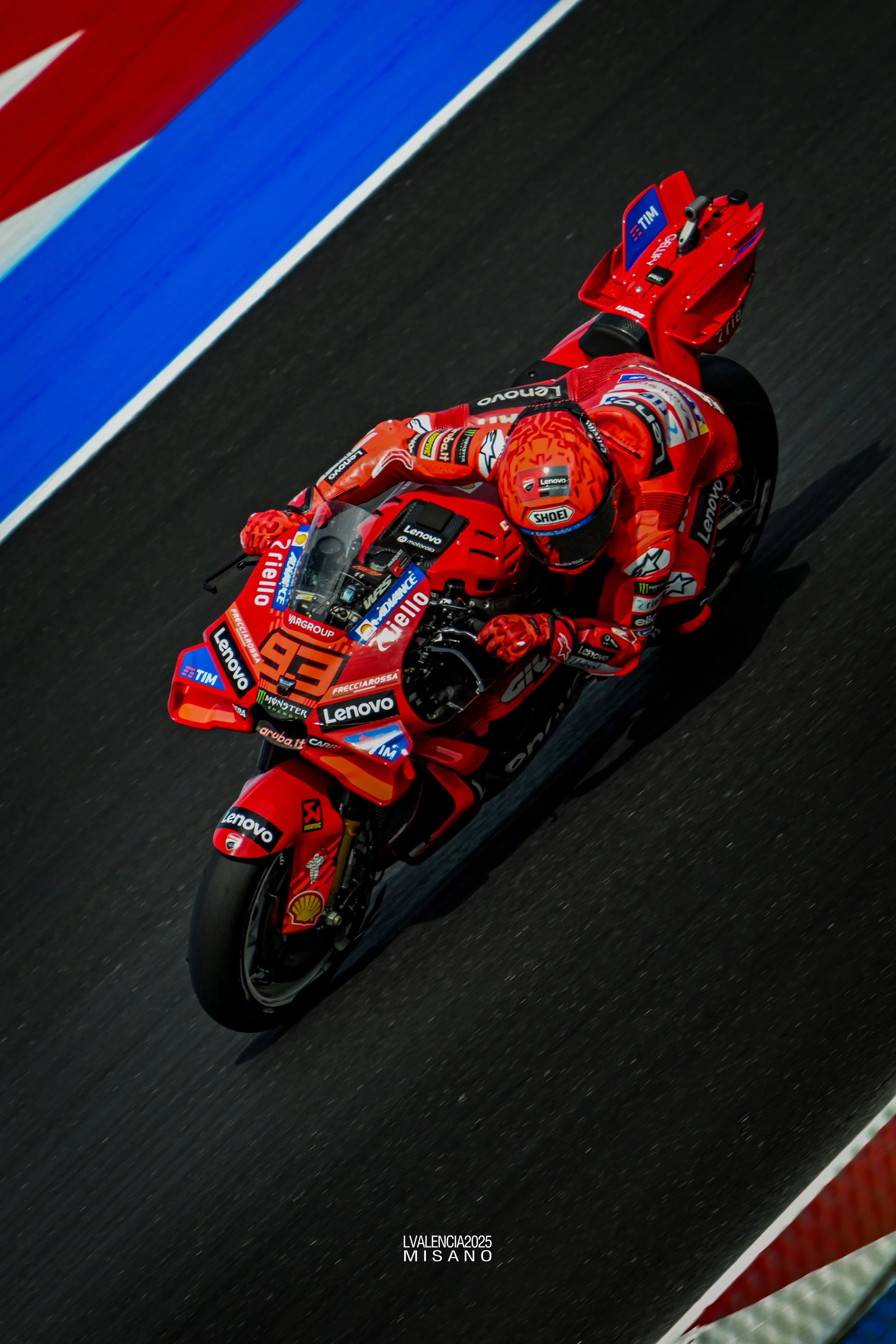 A race motorcycle driven by a rider in a red racing suit and helmet on a dark asphalt track with red, white, and blue painted lines.