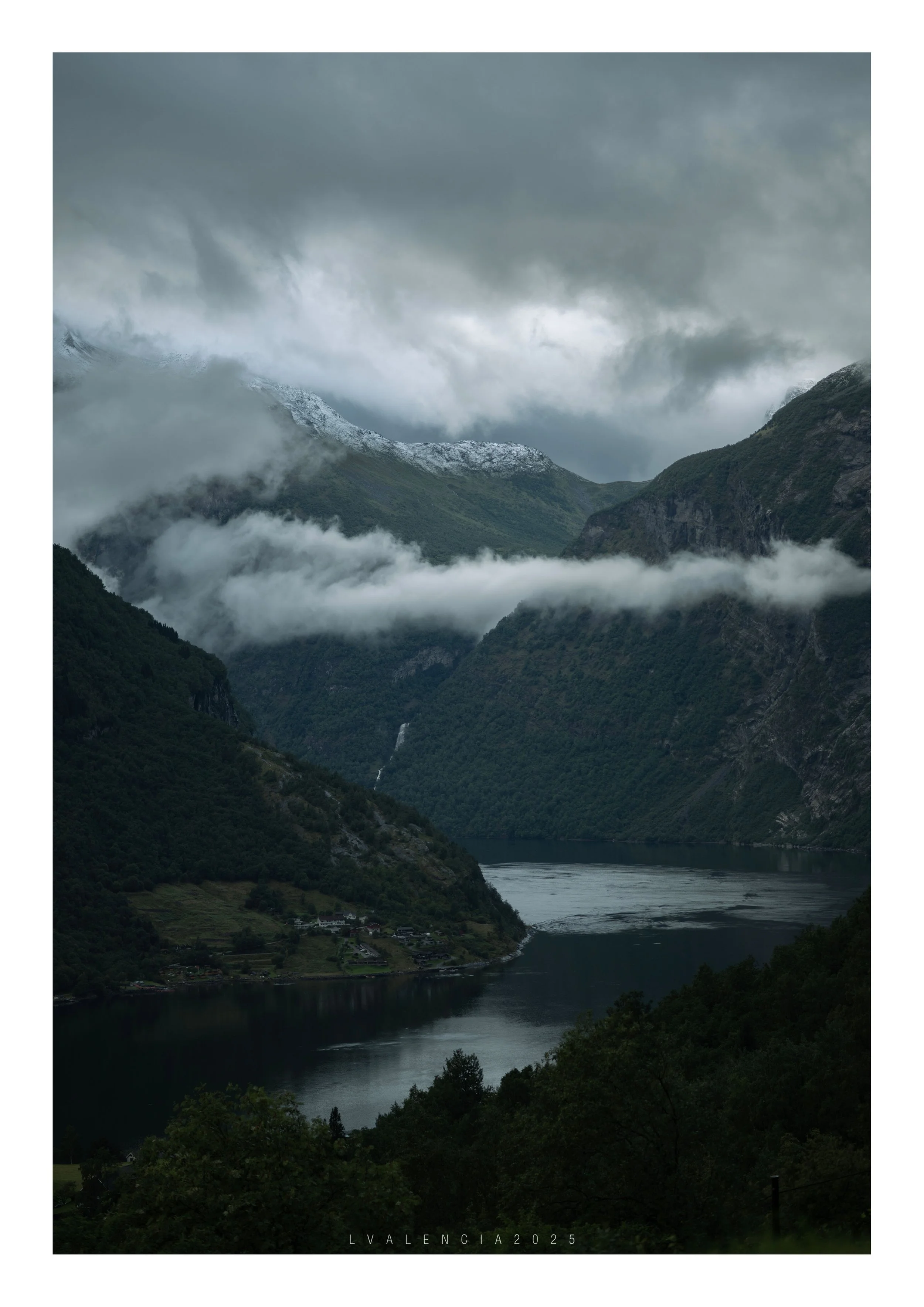 A misty fjord landscape with mountains, some covered in snow, and a river or lake at the bottom. Low clouds partially obscure the mountains.