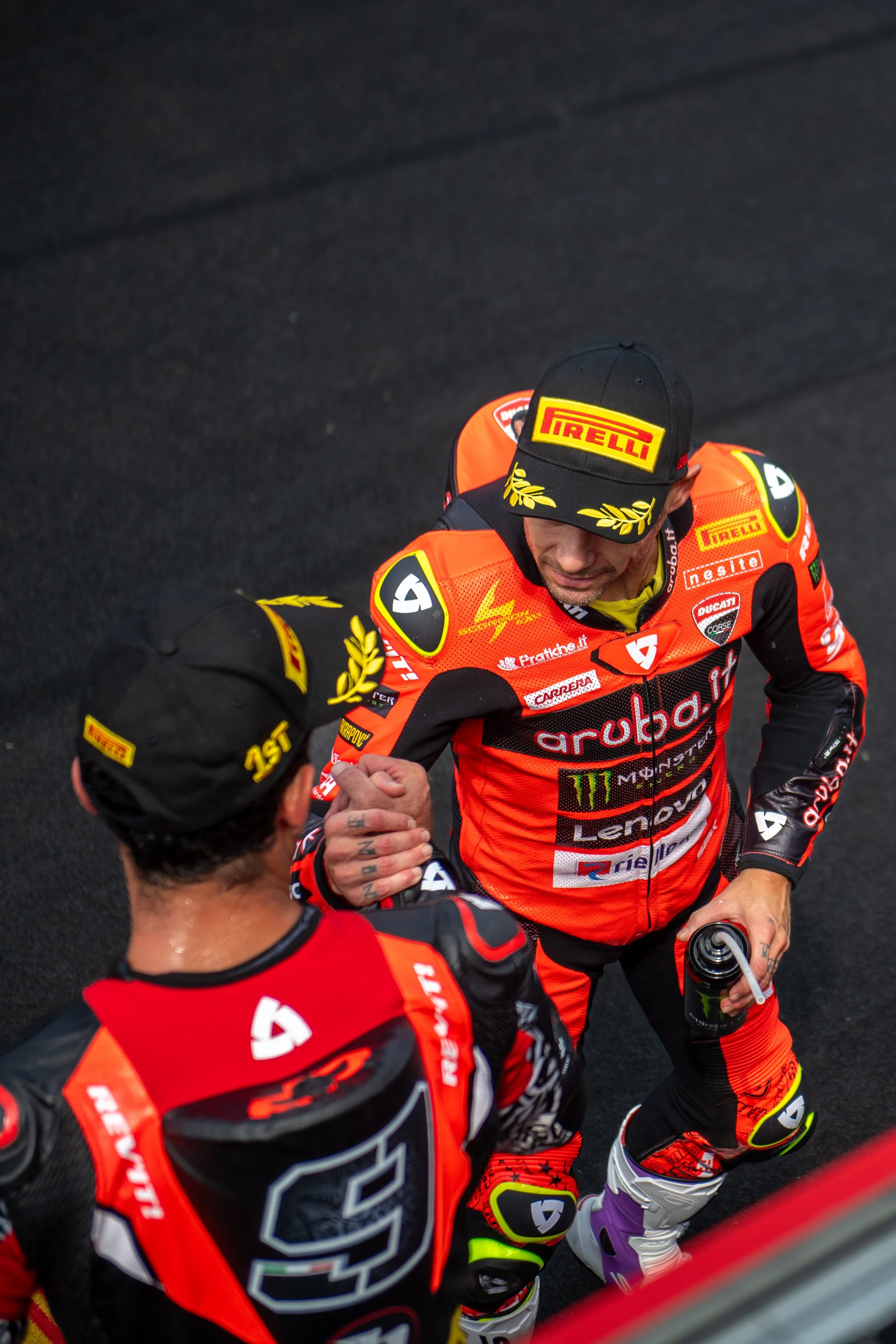 Two race car drivers in racing suits and Pirelli hats shaking hands and greeting each other after a race.