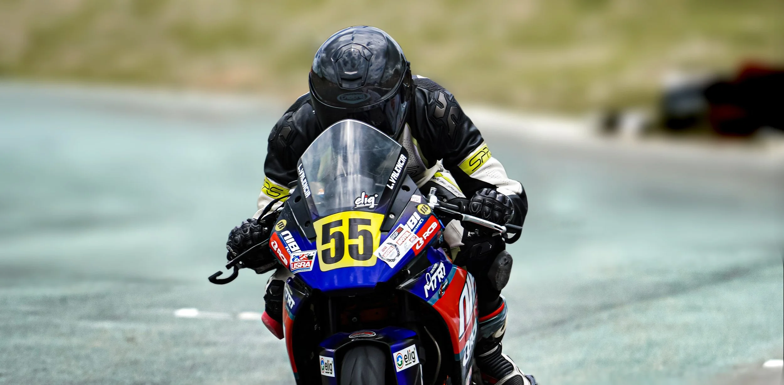 Motorcycle racer wearing black helmet and racing suit, riding a blue, black, red, and yellow motorcycle numbered 55 on a race track. rider name Lawrence Valencia