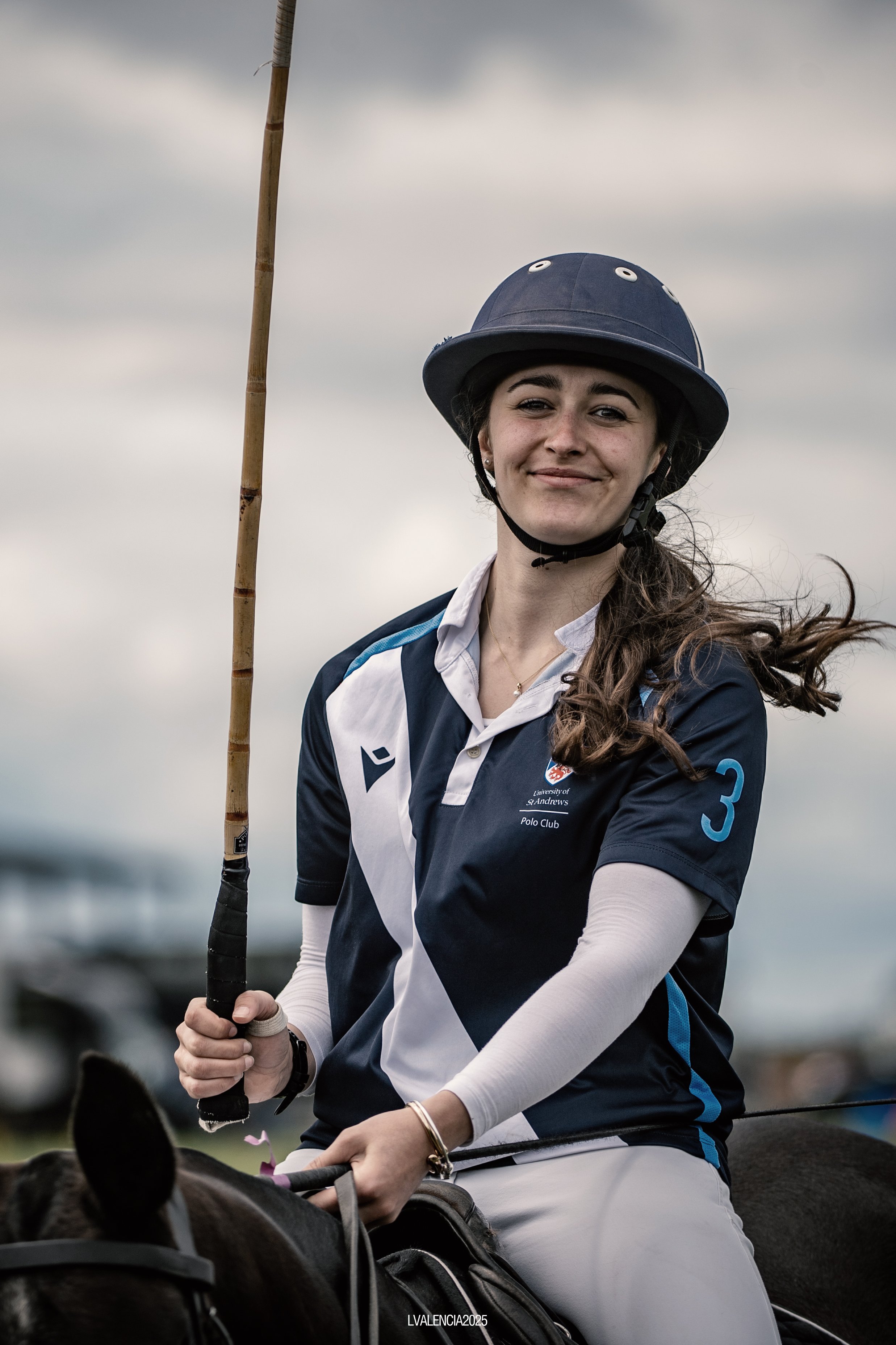 A young woman wearing a navy blue polo shirt with the number 3, a white long-sleeve shirt underneath, and a navy helmet while riding a horse outdoors. She is smiling and holding a polo mallet.