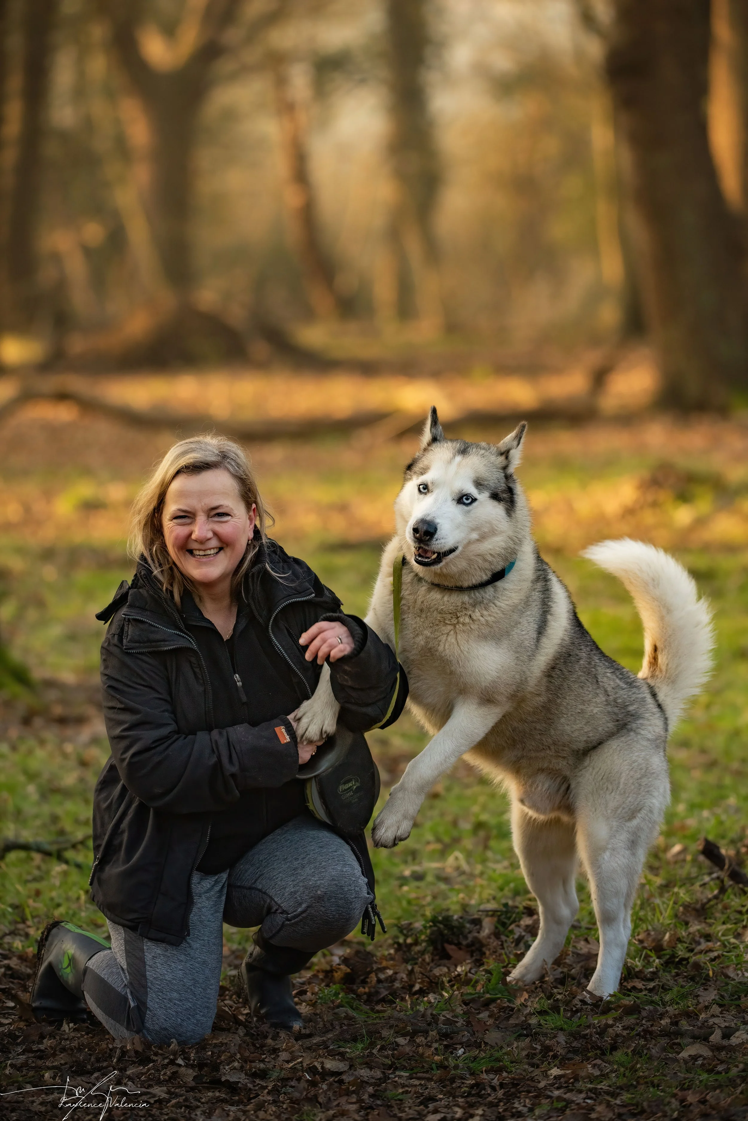 A woman in outdoor clothing kneels on the ground, smiling, holding a Siberian Husky puppy by the paws in a forest setting during autumn.