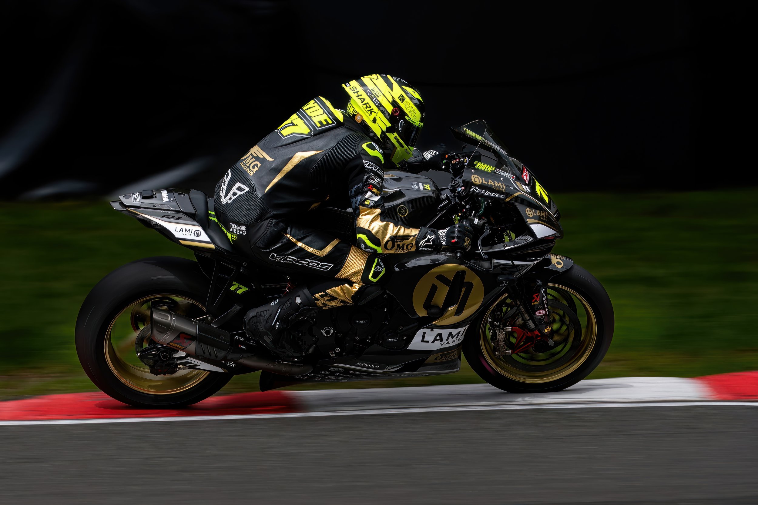 A professional motorcycle racer in black and yellow gear riding a black and gold motorcycle on a racetrack at high speed, with a blurred background and red and white curbing.
