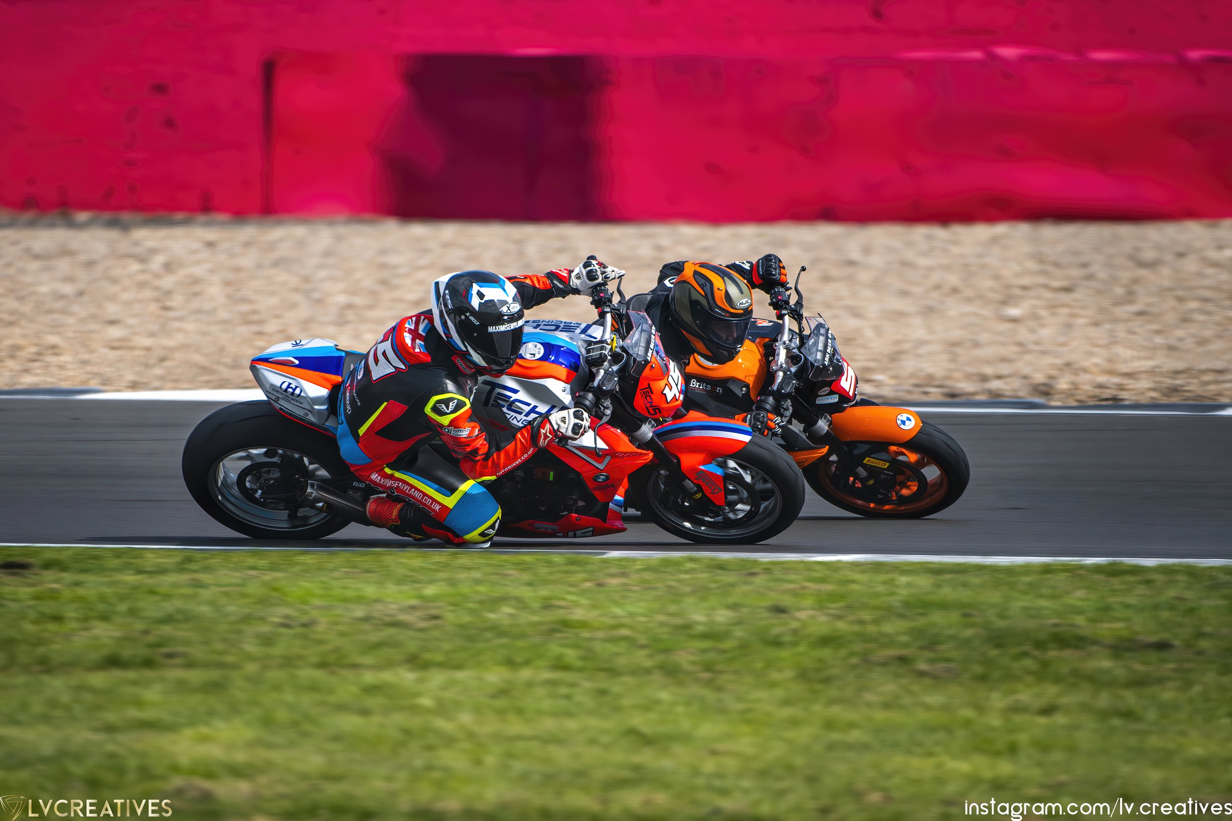 Two motorcycle racers in racing suits and helmets leaning into a turn on a racetrack, with a red barrier in the background.