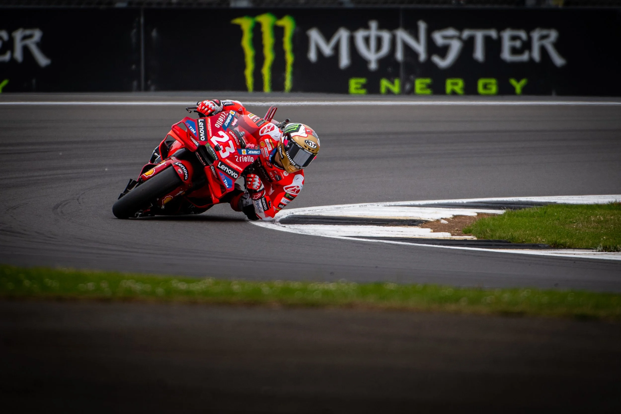 A professional motorcycle racer in red gear leaning into a sharp turn on a race track, with a Monster Energy advertisement in the background.