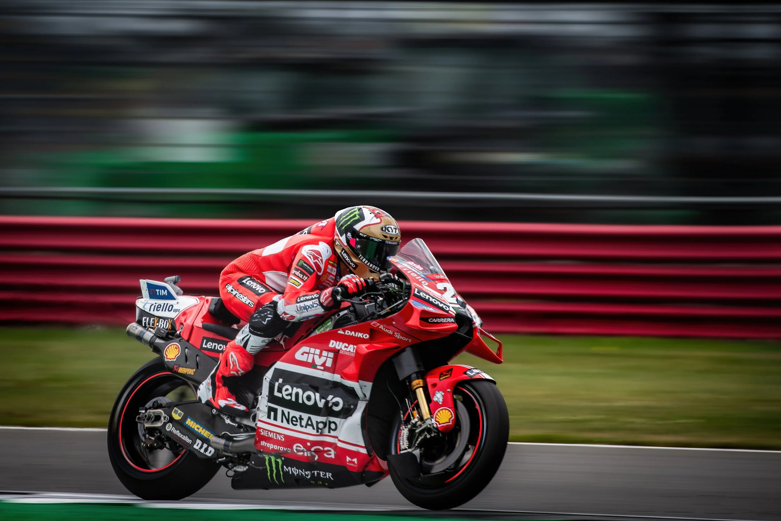 A professional motorcycle racer in red and black gear riding a red Ducati motorcycle on a racetrack, with a blurred background indicating high speed.