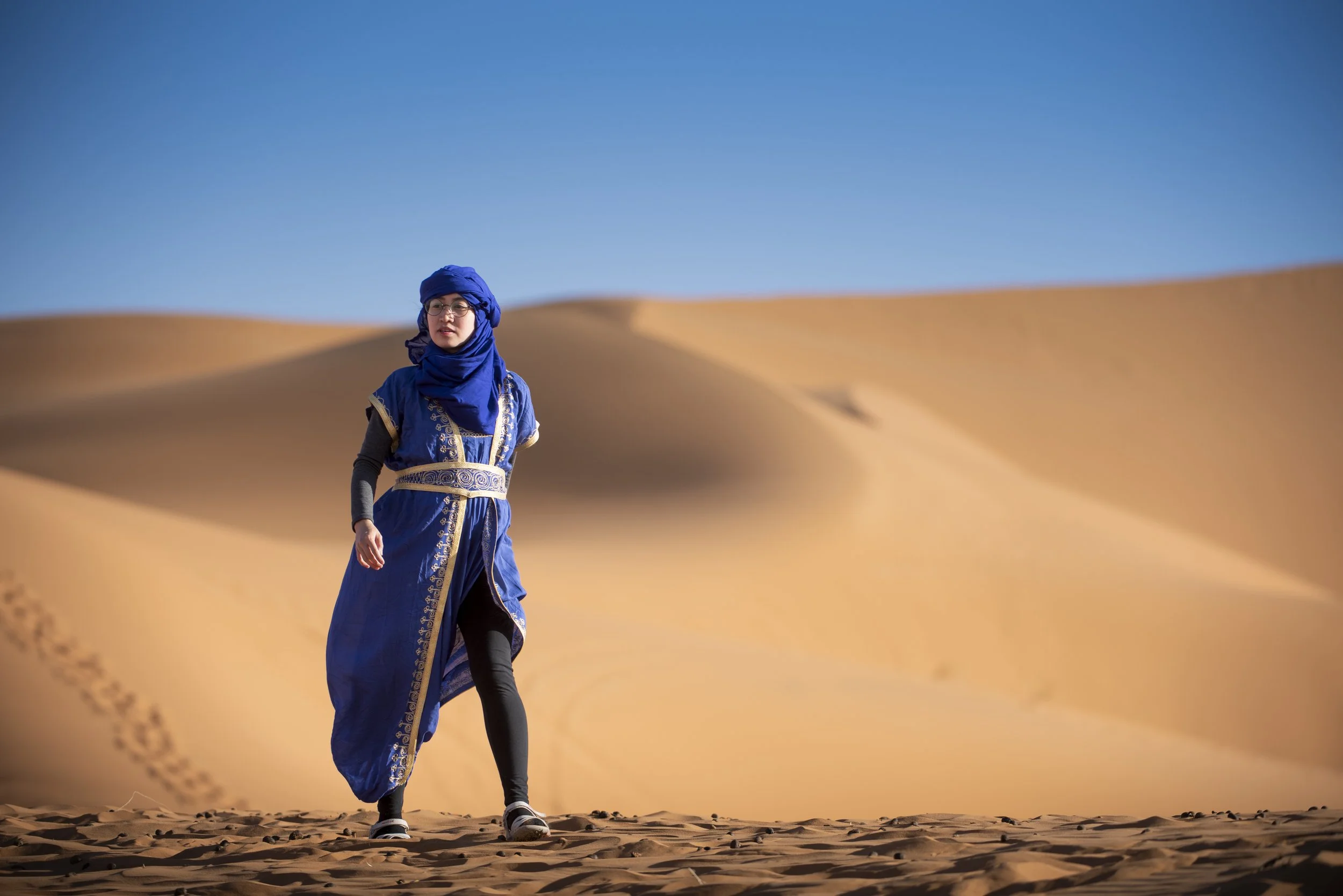 Person dressed in blue traditional clothing walking in desert with sand dunes and clear blue sky.