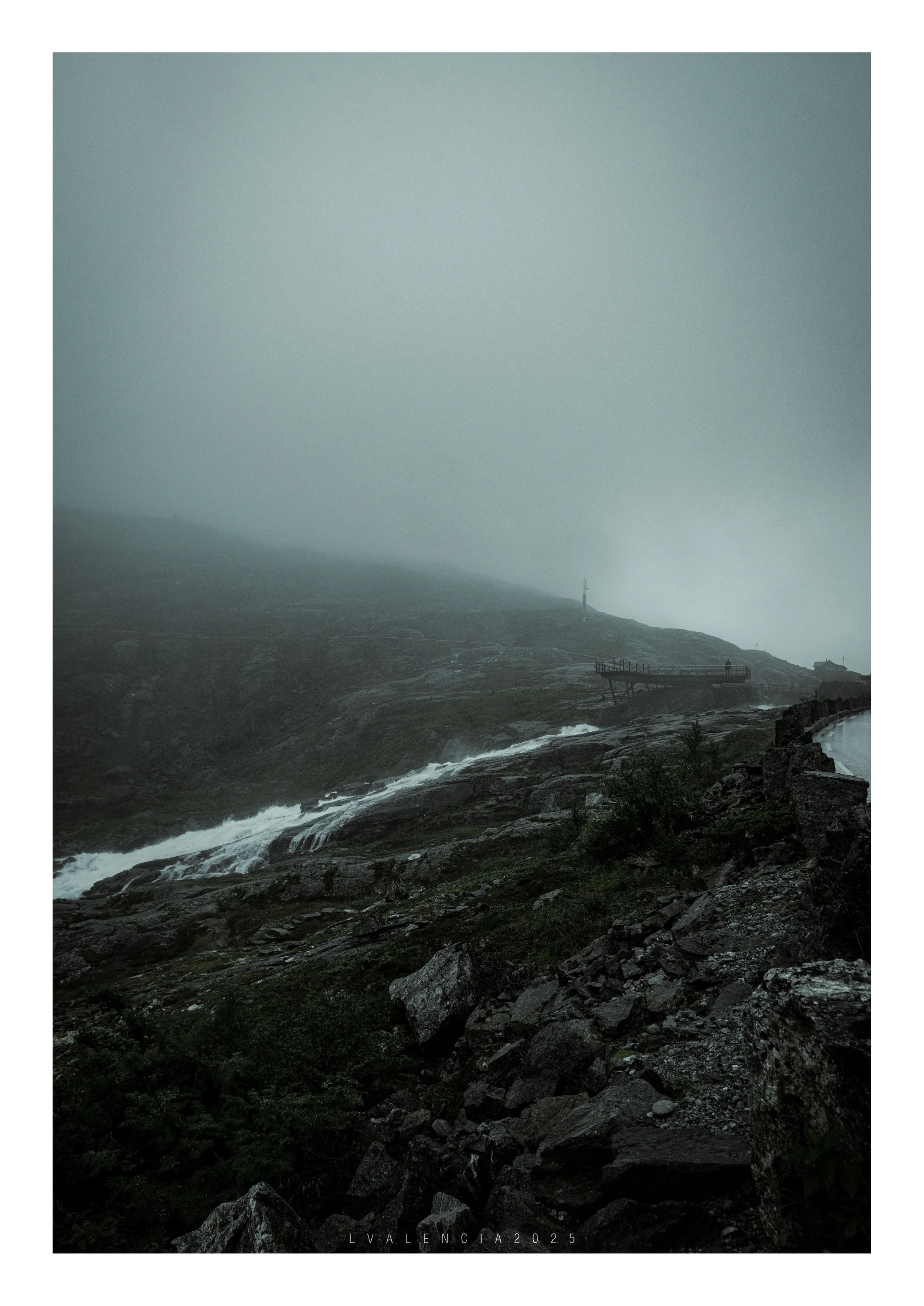 A foggy and rainy coastal landscape with rocks, a flowing stream, a winding road, and a small structure. The sky is overcast, and the scene has a moody atmosphere.
