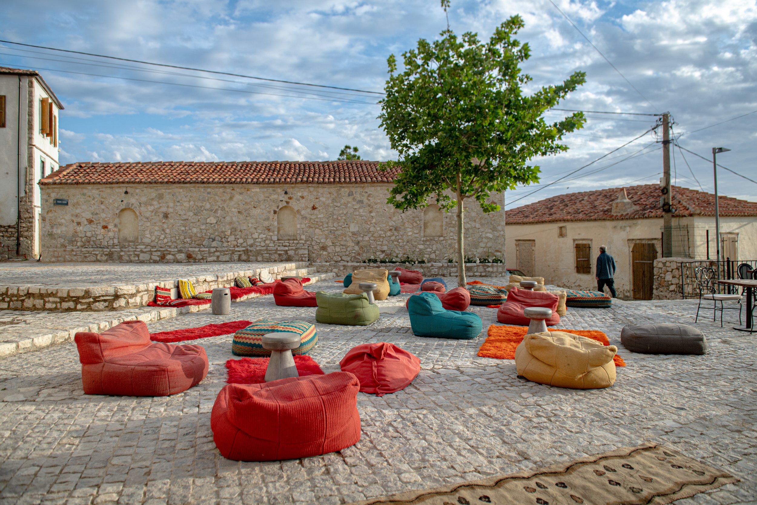 An outdoor courtyard with colorful bean bags and small stools, stone pavement, a tree in the center, stone walls, and a cloudy sky overhead.