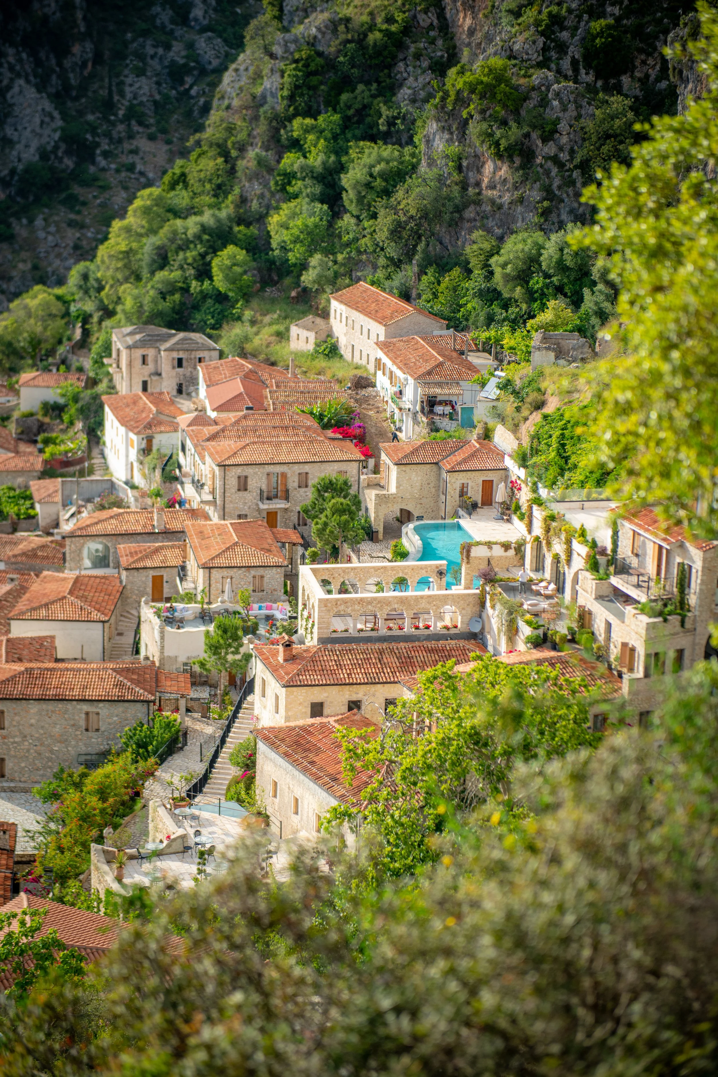 A picturesque hillside village with stone buildings, red-tiled roofs, lush greenery, and a swimming pool, surrounded by forested cliffs.