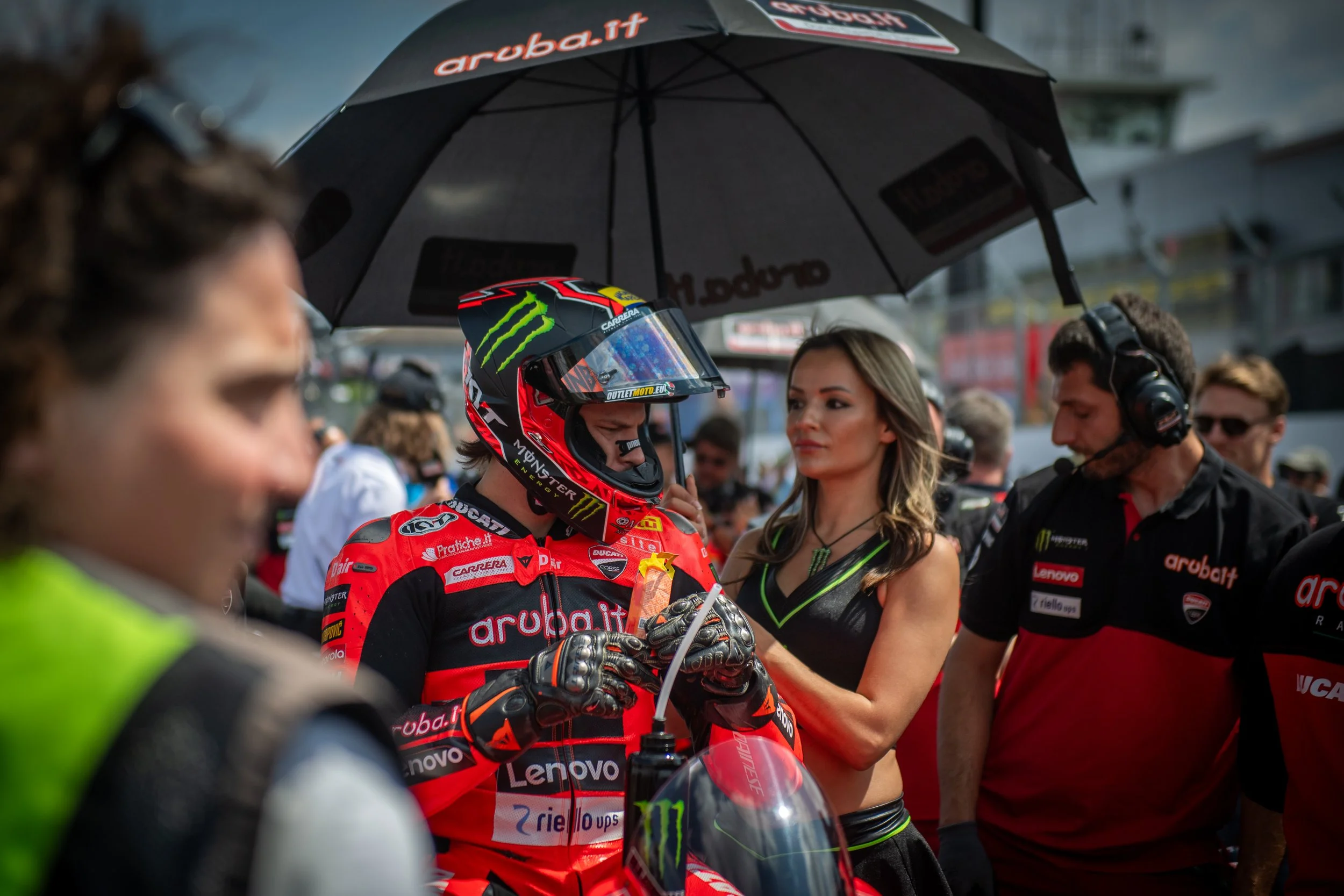 A motorcycle racer in red and black gear standing in a pit area, with a helmet and team members around him, one holding an umbrella over his head during a race event.