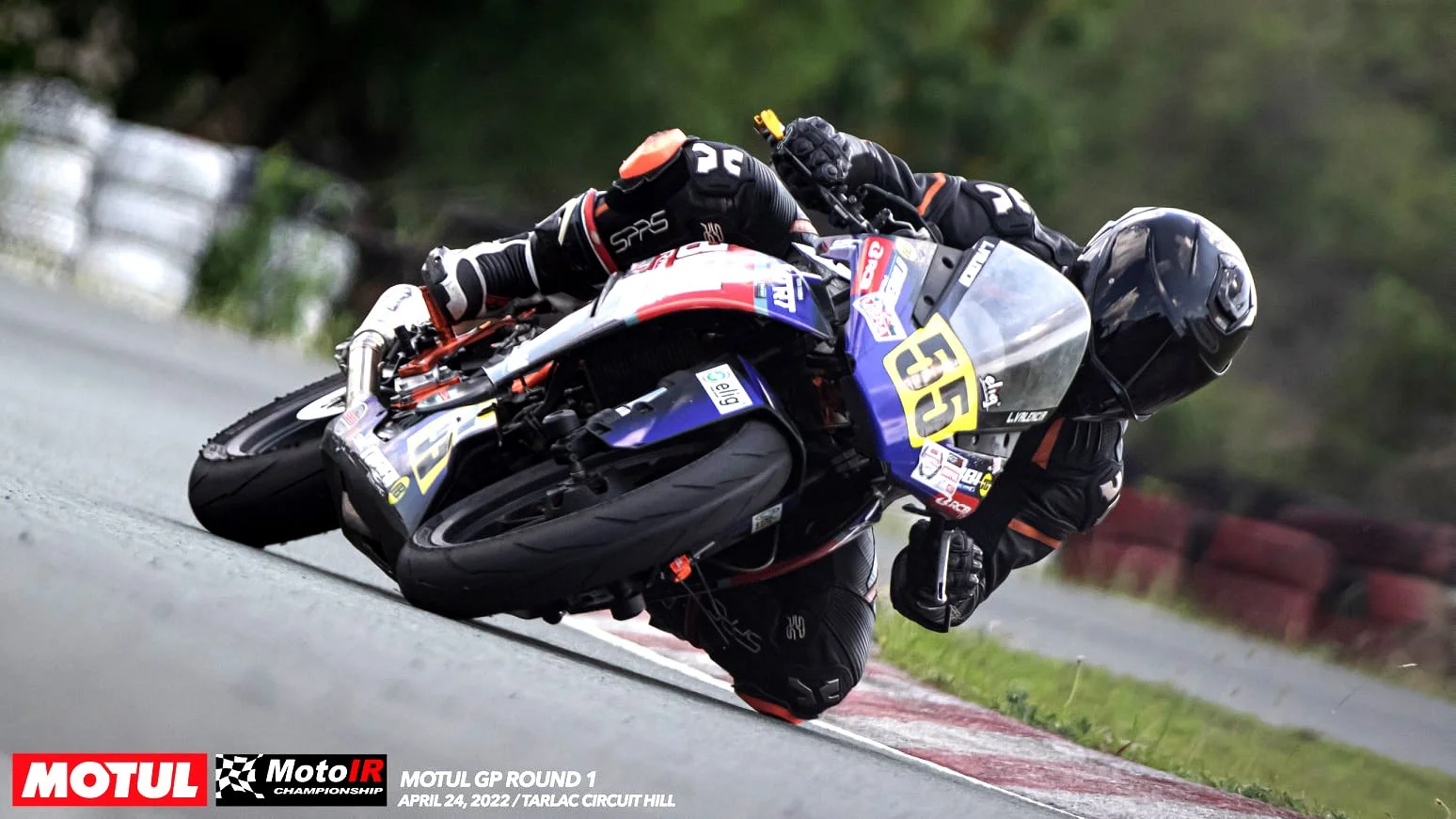 Motorcycle racer leaning into a turn on a race track, wearing full racing gear and helmet, with a blurred green background and race track facilities. Branding and event details are visible at the bottom. rider name Lawrence Valencia