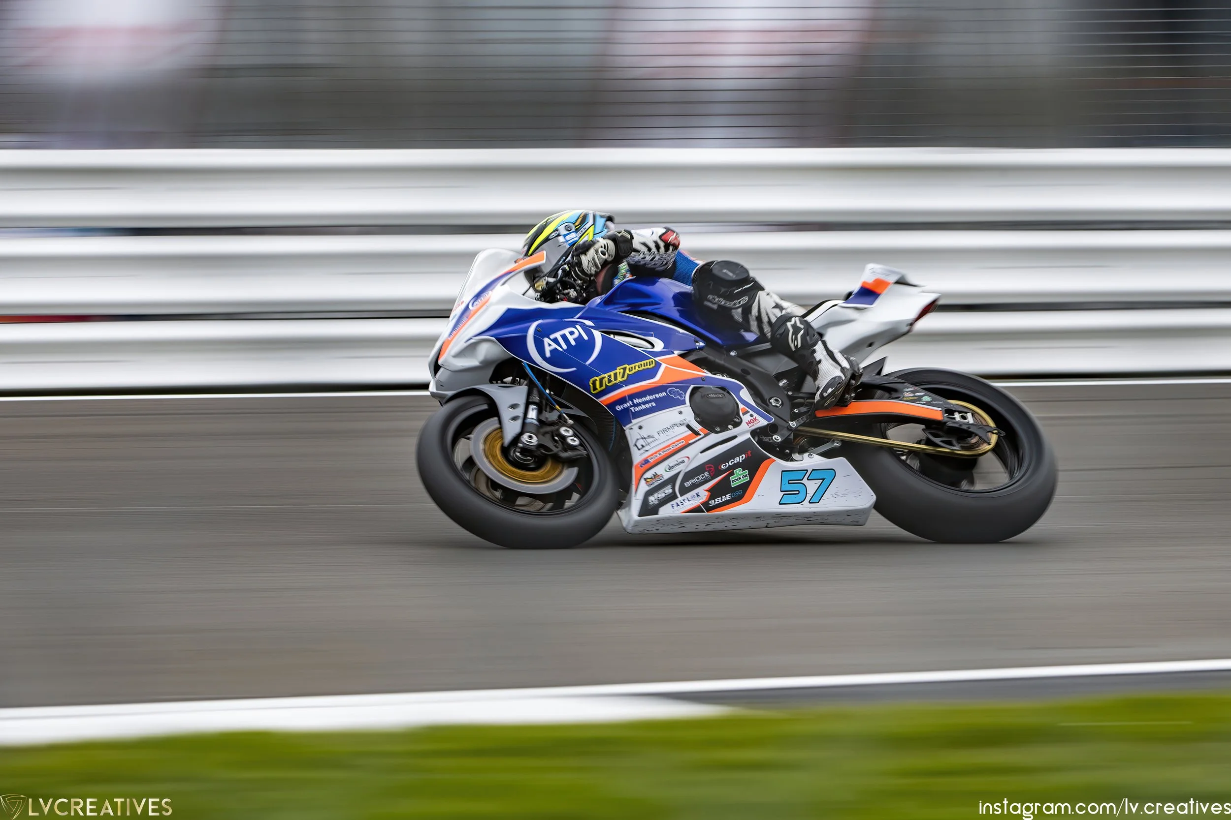 Motorcycle racer leaning into a turn on a race track, wearing a full racing suit and helmet, with the number 57 displayed on the bike.