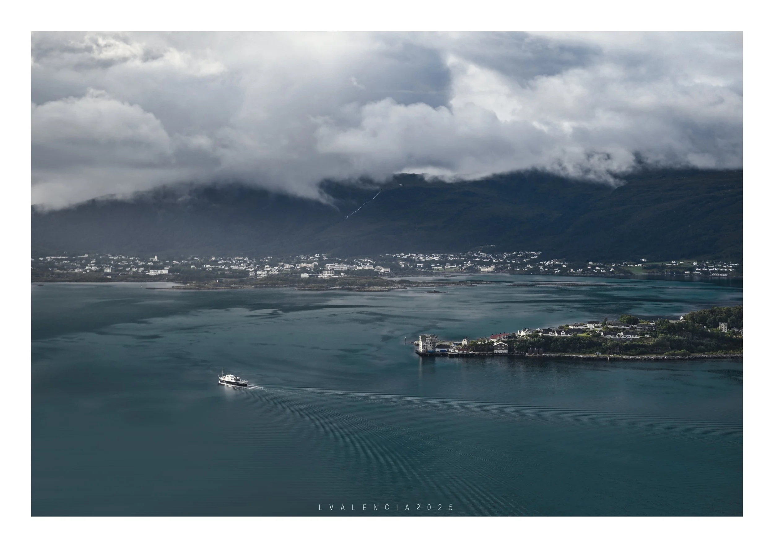 A harbor with a boat creating ripples in the water, surrounded by buildings, and mountains with clouds in the background.