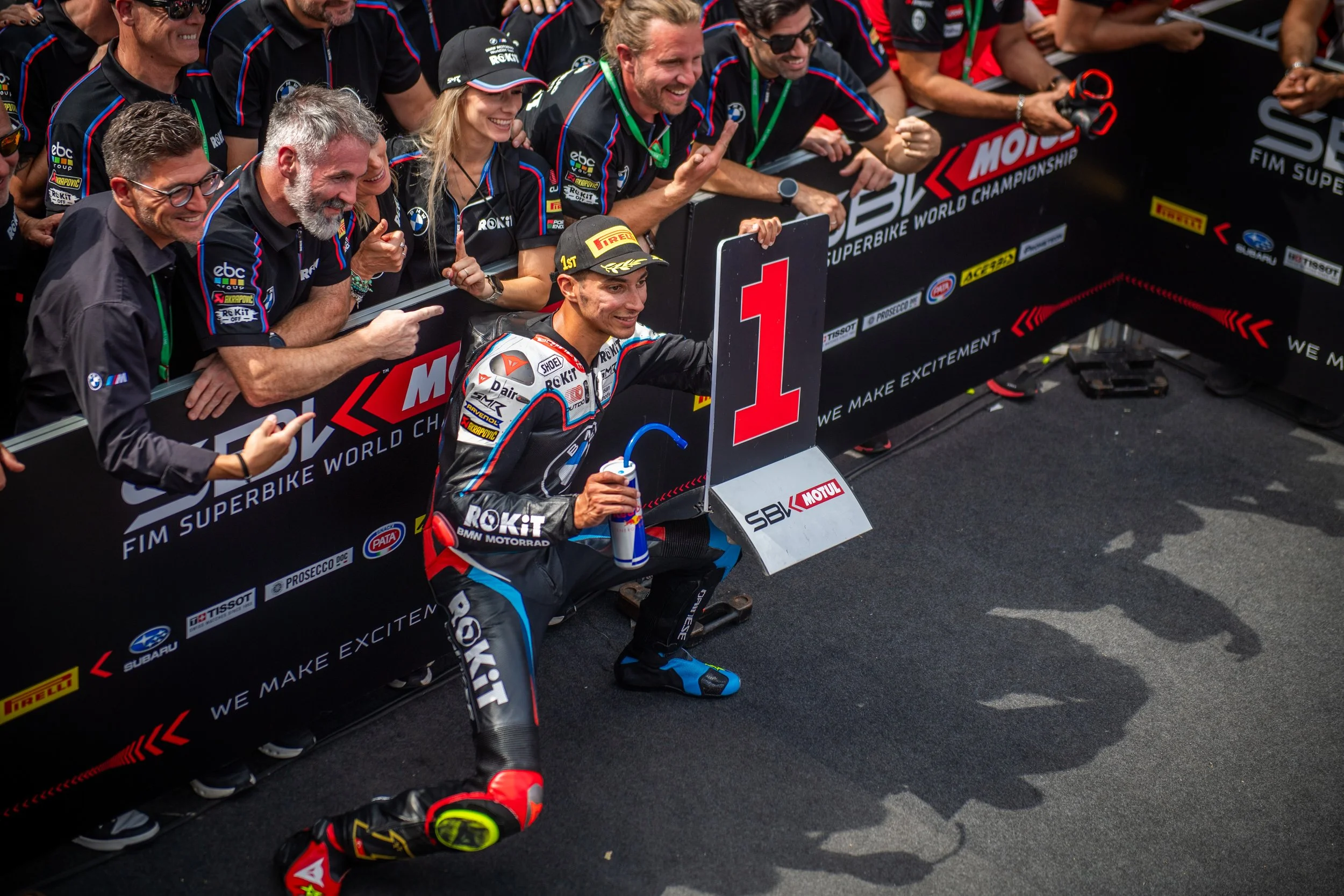 Motorcycle racer kneeling next to a large 1 sign, smiling, holding a drink and a spray, surrounded by team members celebrating at the finish line of a motorcycle race.