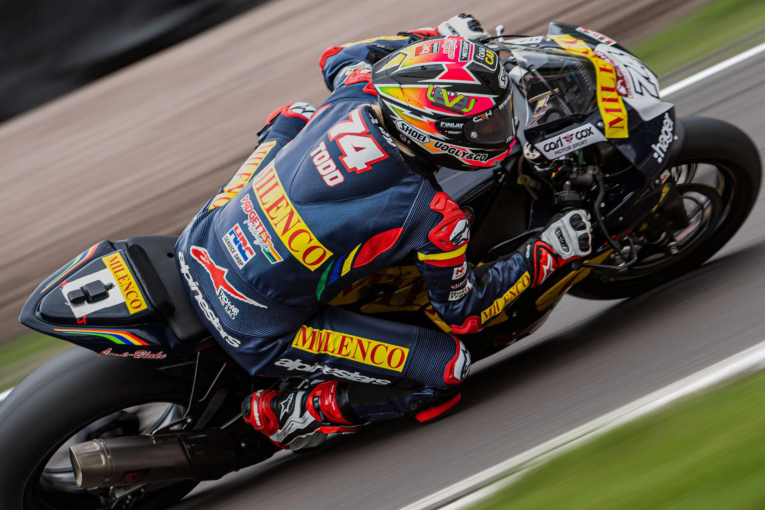 Motorcycle racer leaning into a turn, wearing a blue racing suit with red, yellow, and green accents, and a colorful helmet, on a black sport motorcycle with sponsor decals.