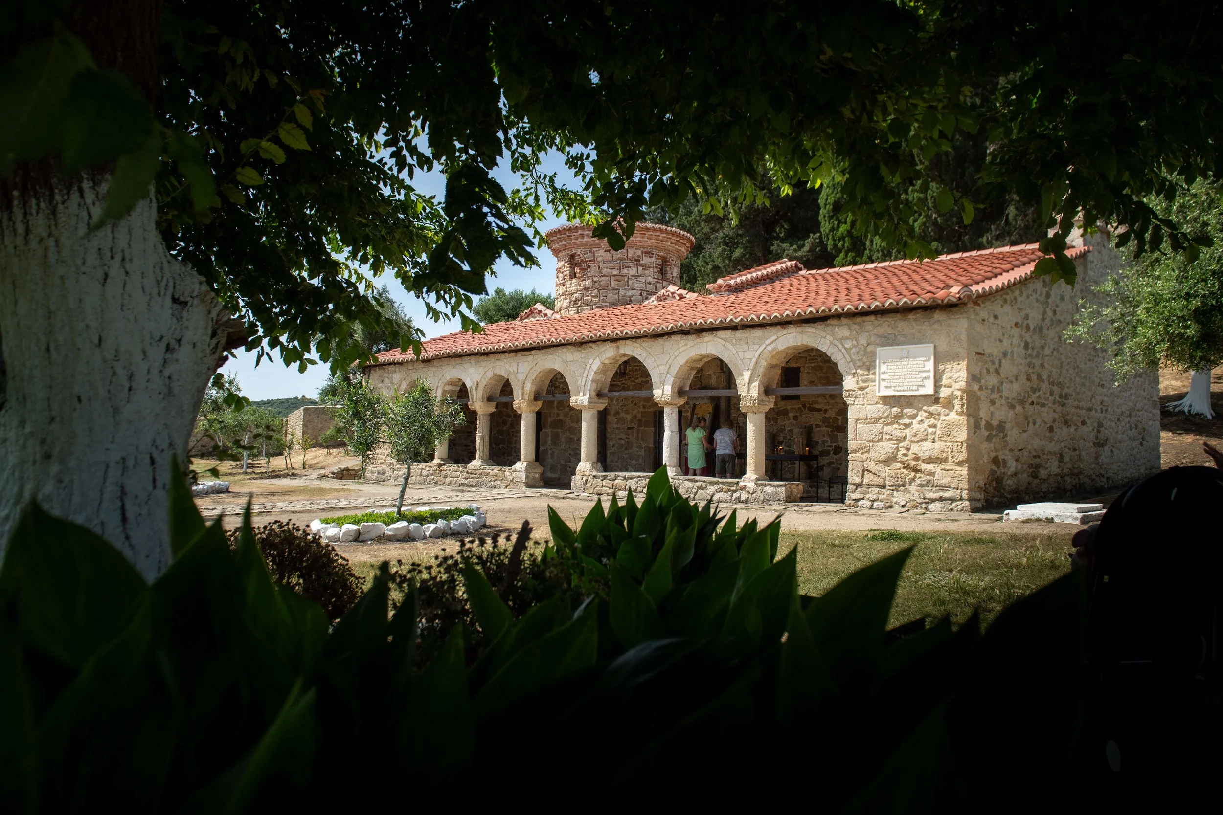 A small historic stone building with a red-tiled roof, arched portico, and tower, surrounded by trees and bushes, with visitors near the entrance.