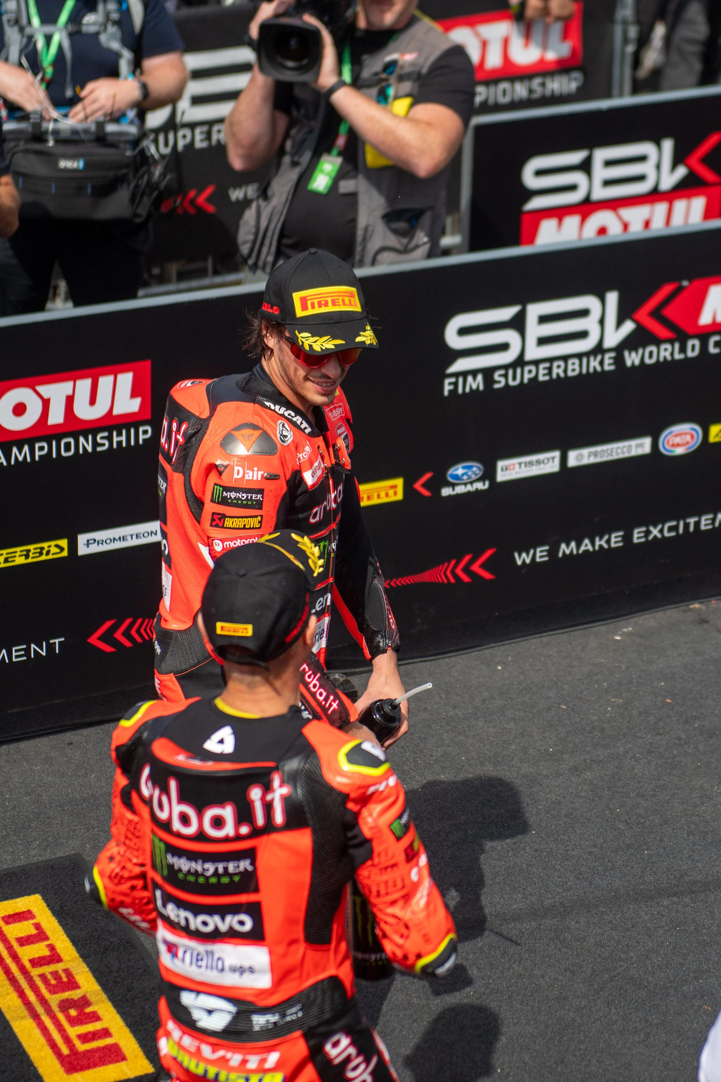 Two motorcycle racers in racing suits and black caps, one with a bottle, standing in front of a sponsor backdrop at the SBK Superbike World Championship event.
