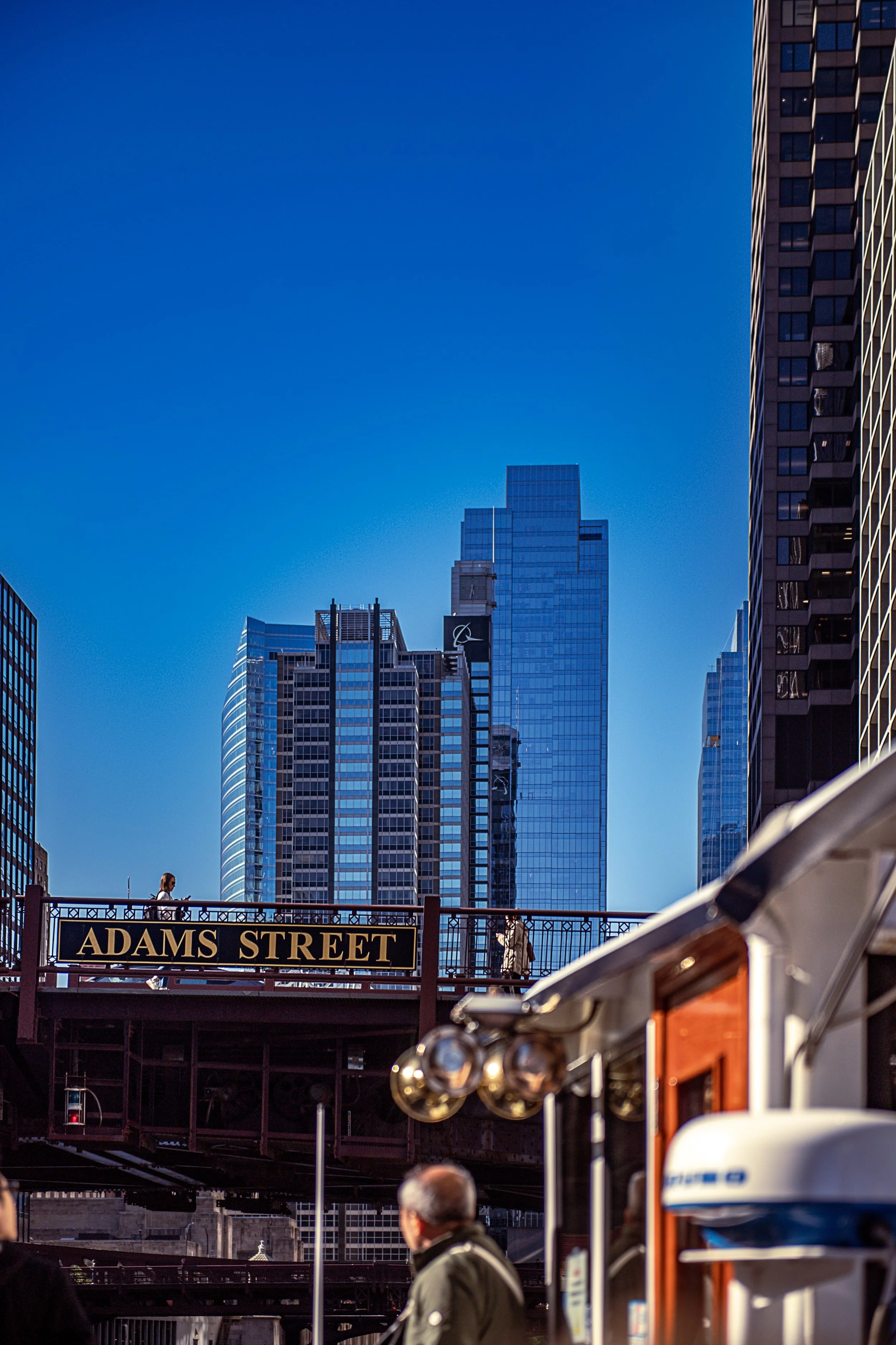 A city scene on a sunny day with tall skyscrapers, a bridge labeled 'Adams Street,' pedestrians, and a boat in the foreground.