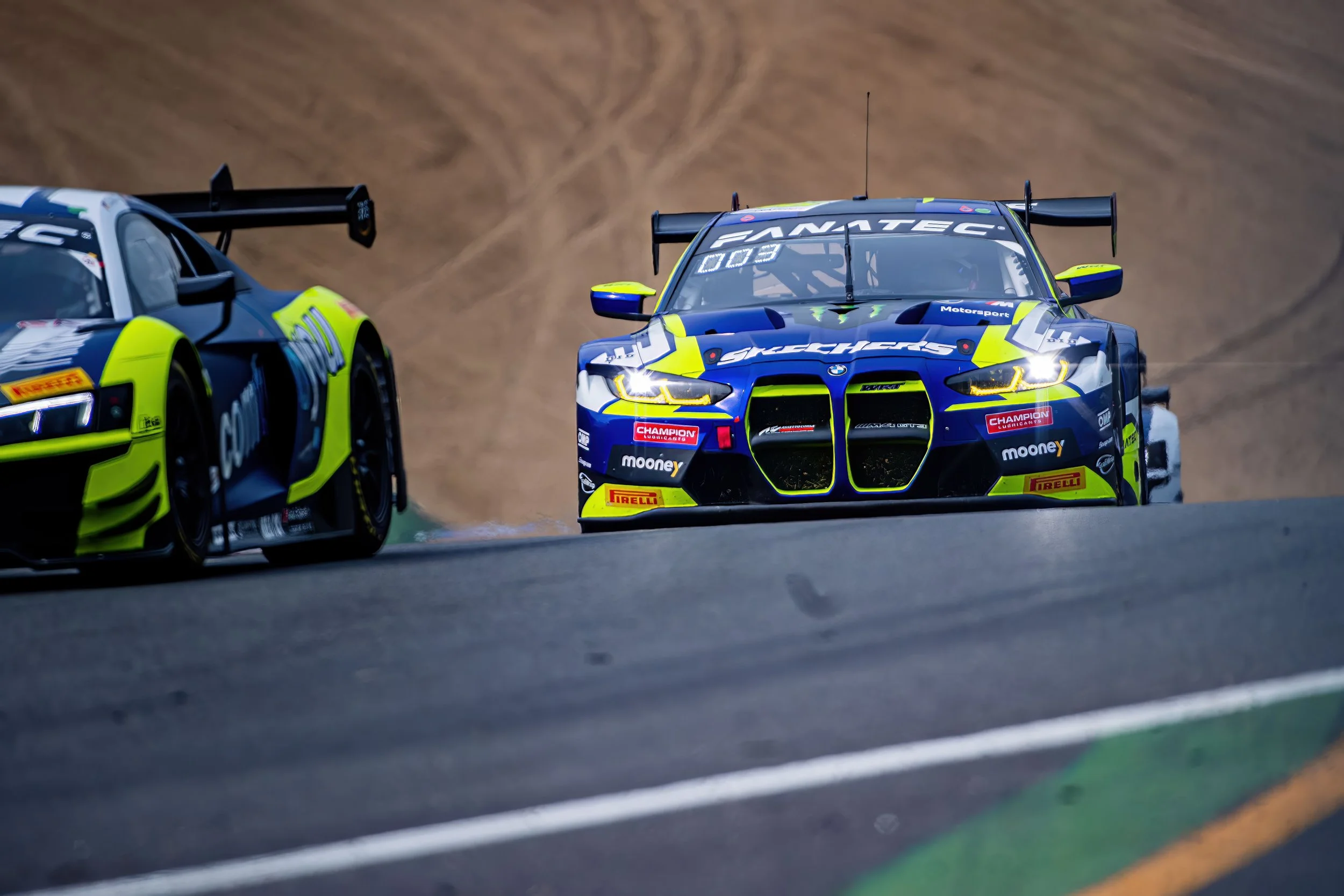 Two race cars on a track, with one BMW M4 GT3 in front and an Audi R8 LMS behind, racing on a curved section of the track with brown and green surroundings.
