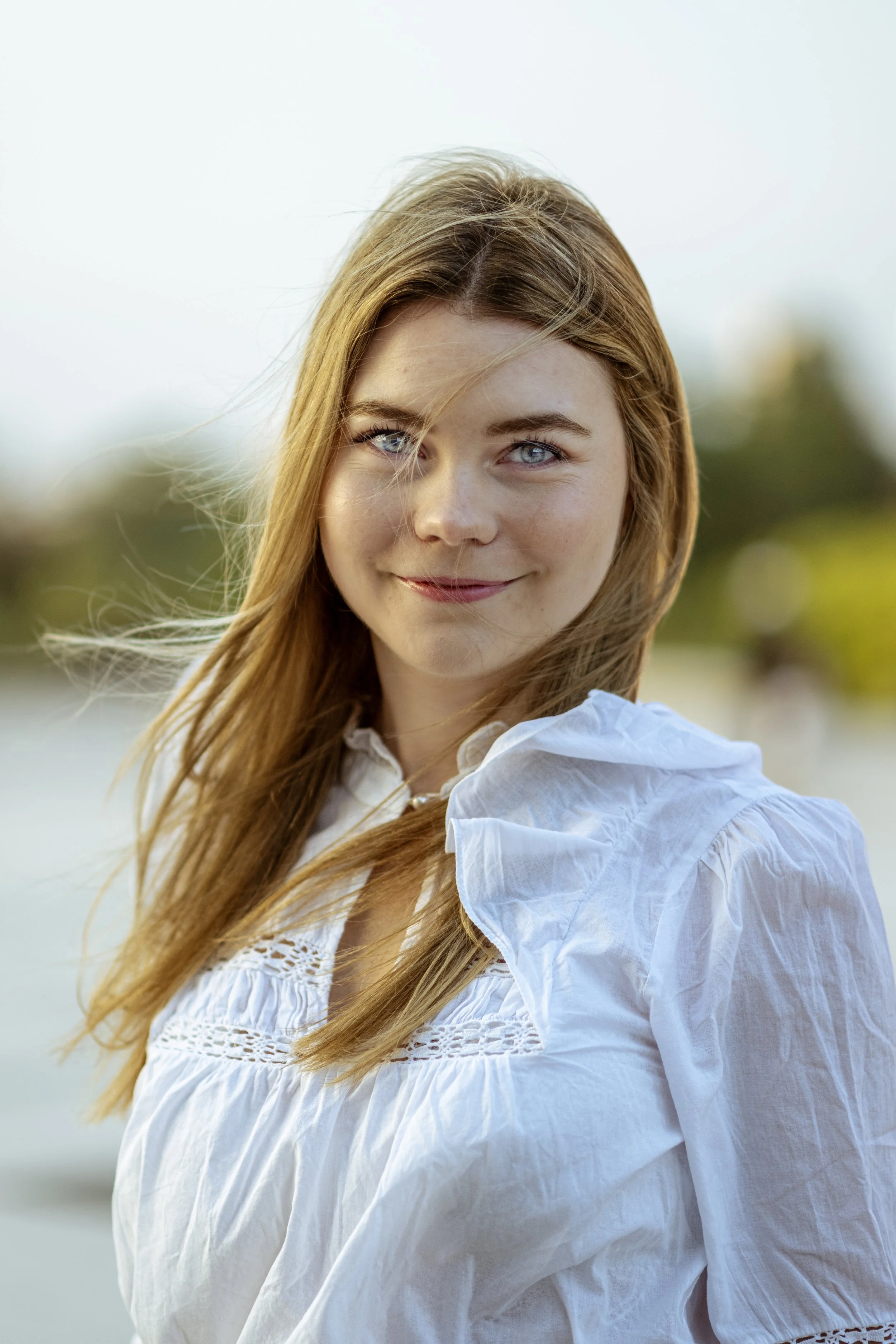 A young woman with long, light brown hair and blue eyes smiling outdoors in natural light, wearing a white blouse.