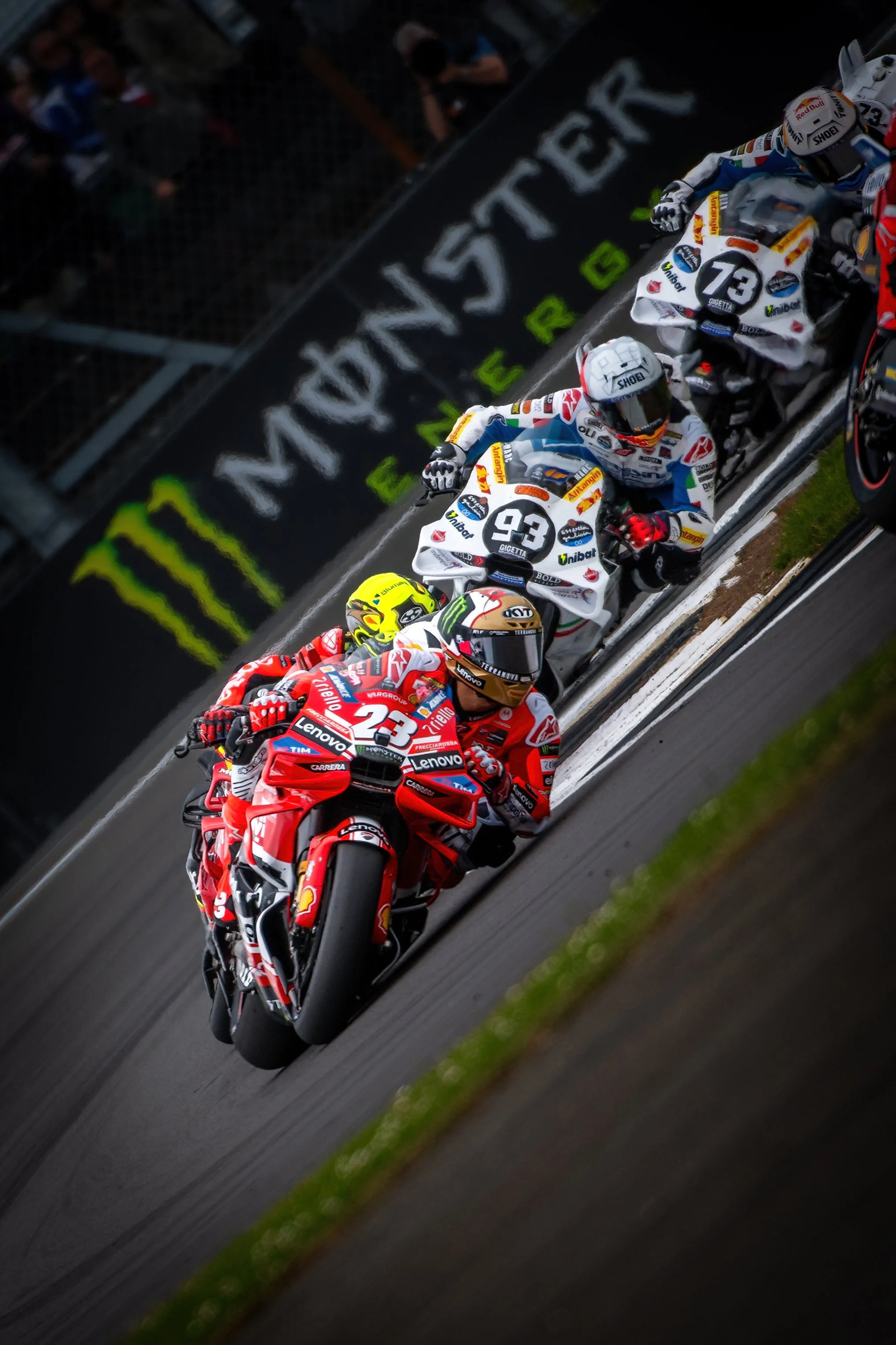 Motorcycle racers lean into a turn during a race, with the leading rider wearing a red suit and helmet, followed by riders in various colored suits and helmets, on a racetrack with advertising banners in the background.