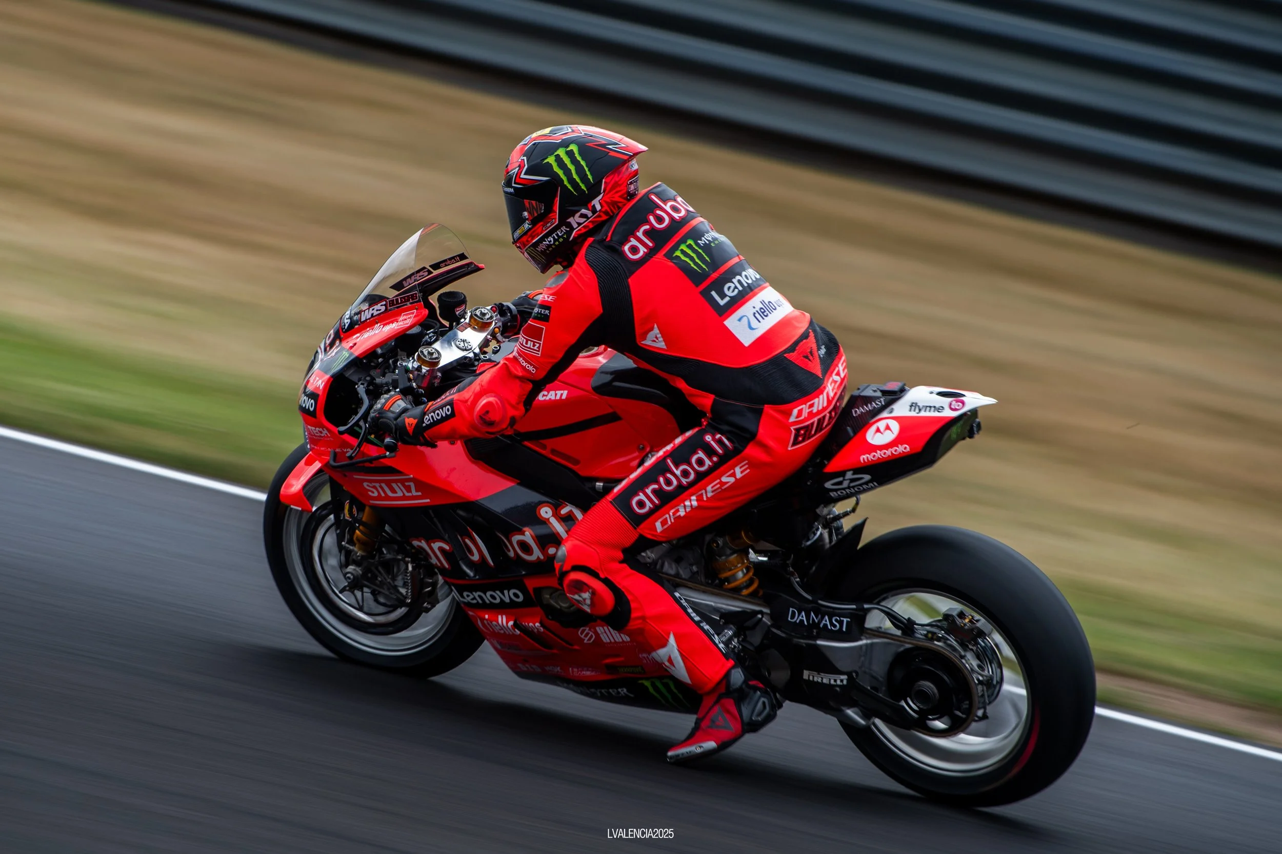 A motorcycle racer in red racing suit and helmet riding a red and black racing motorcycle on a track, with motion blur background.