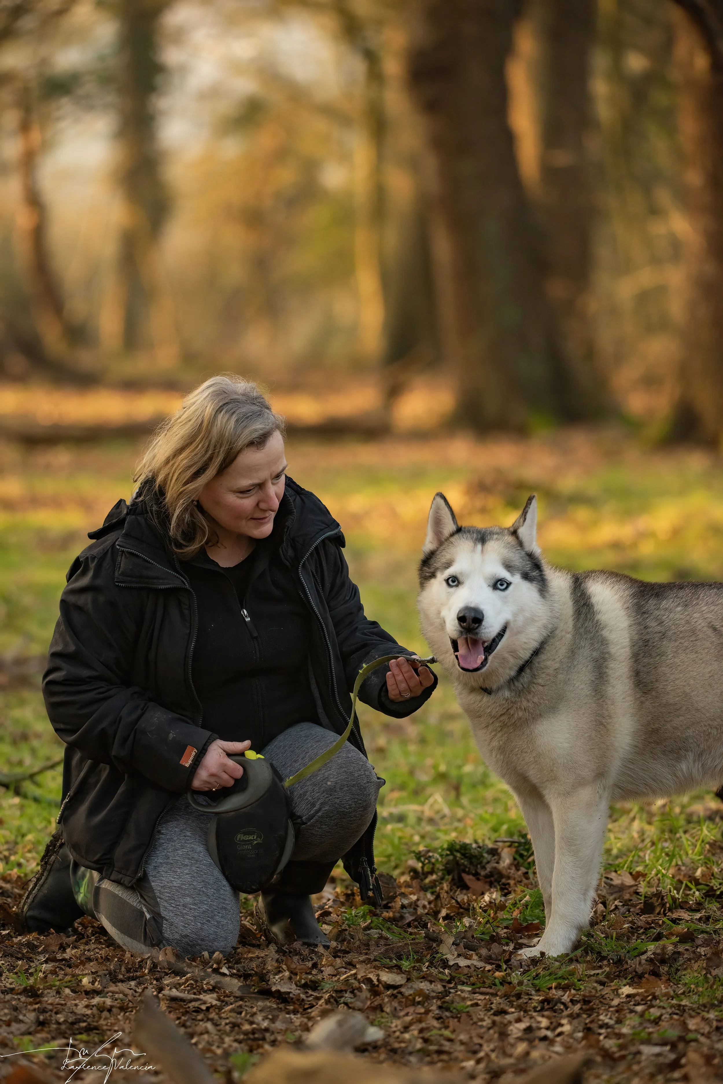 A woman kneeling on the ground in a forest, holding a leash attached to a Siberian Husky with blue eyes, both appearing happy.