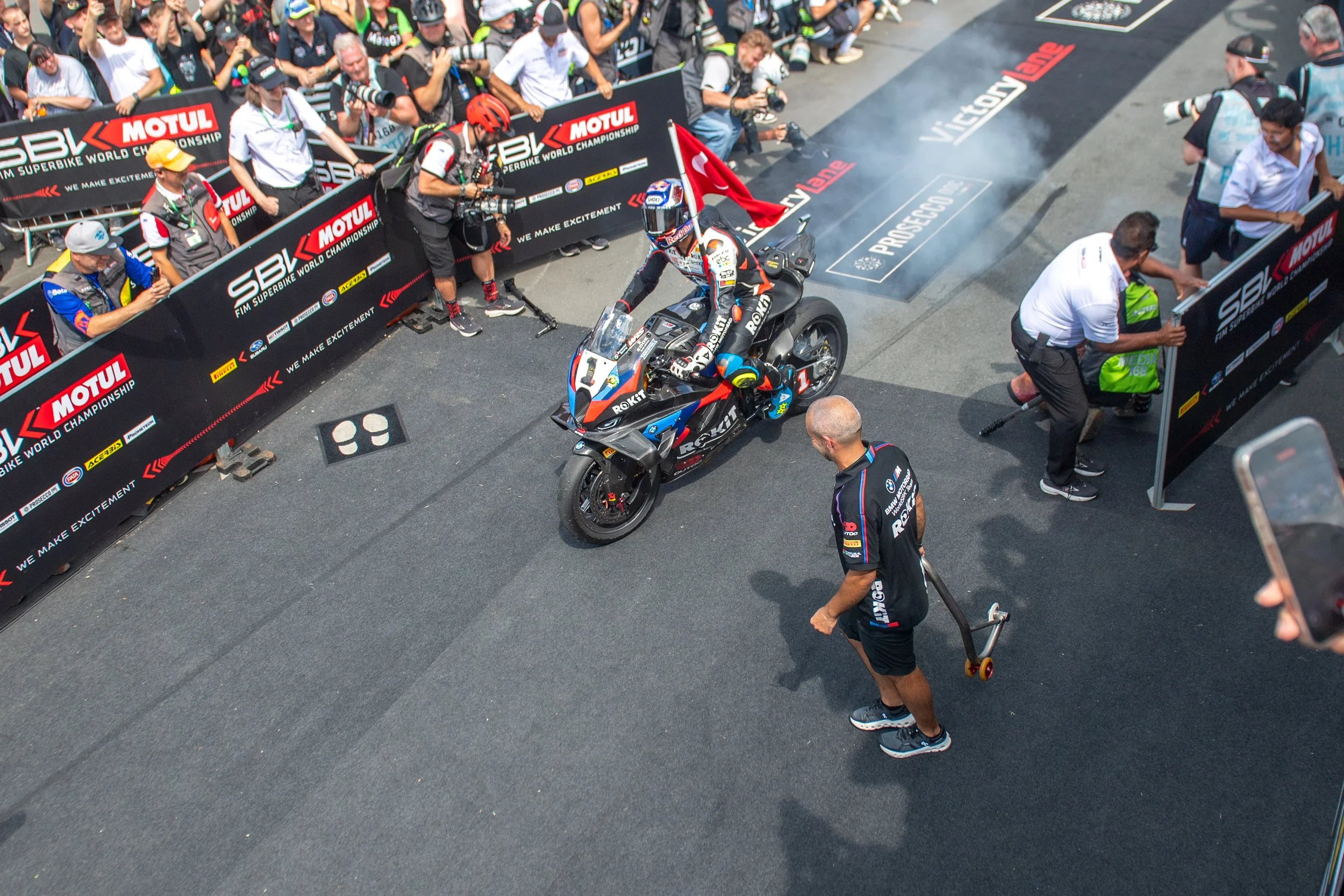 A motorcycle rider, clad in racing gear, sits on a racing motorcycle in the pit lane of a motorsport event. The scene is surrounded by team members and spectators, with banners and advertisements visible, during daytime.